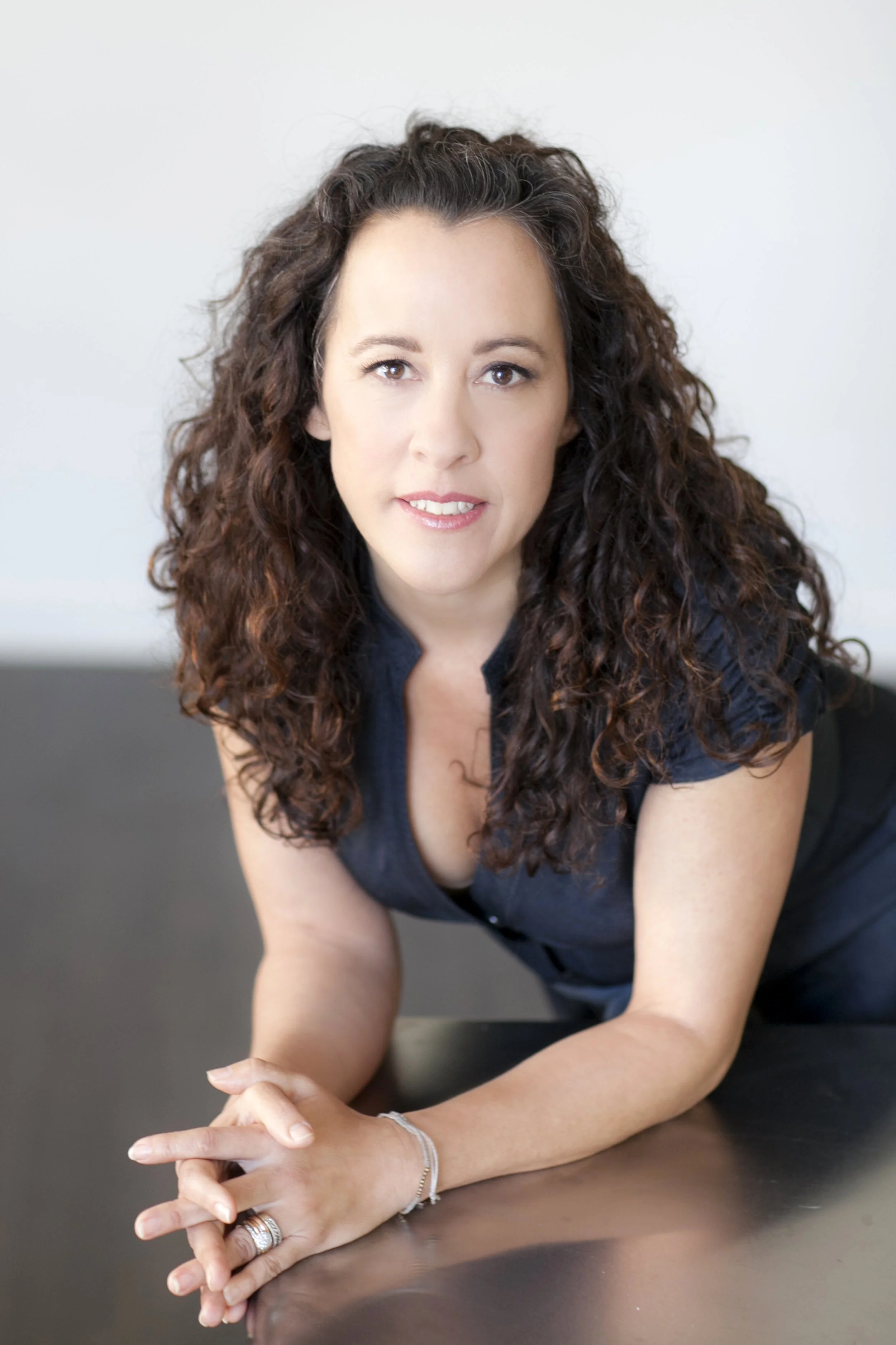 A woman with curly brown hair leaning on a dark table, wearing a dark top, looking at the camera.