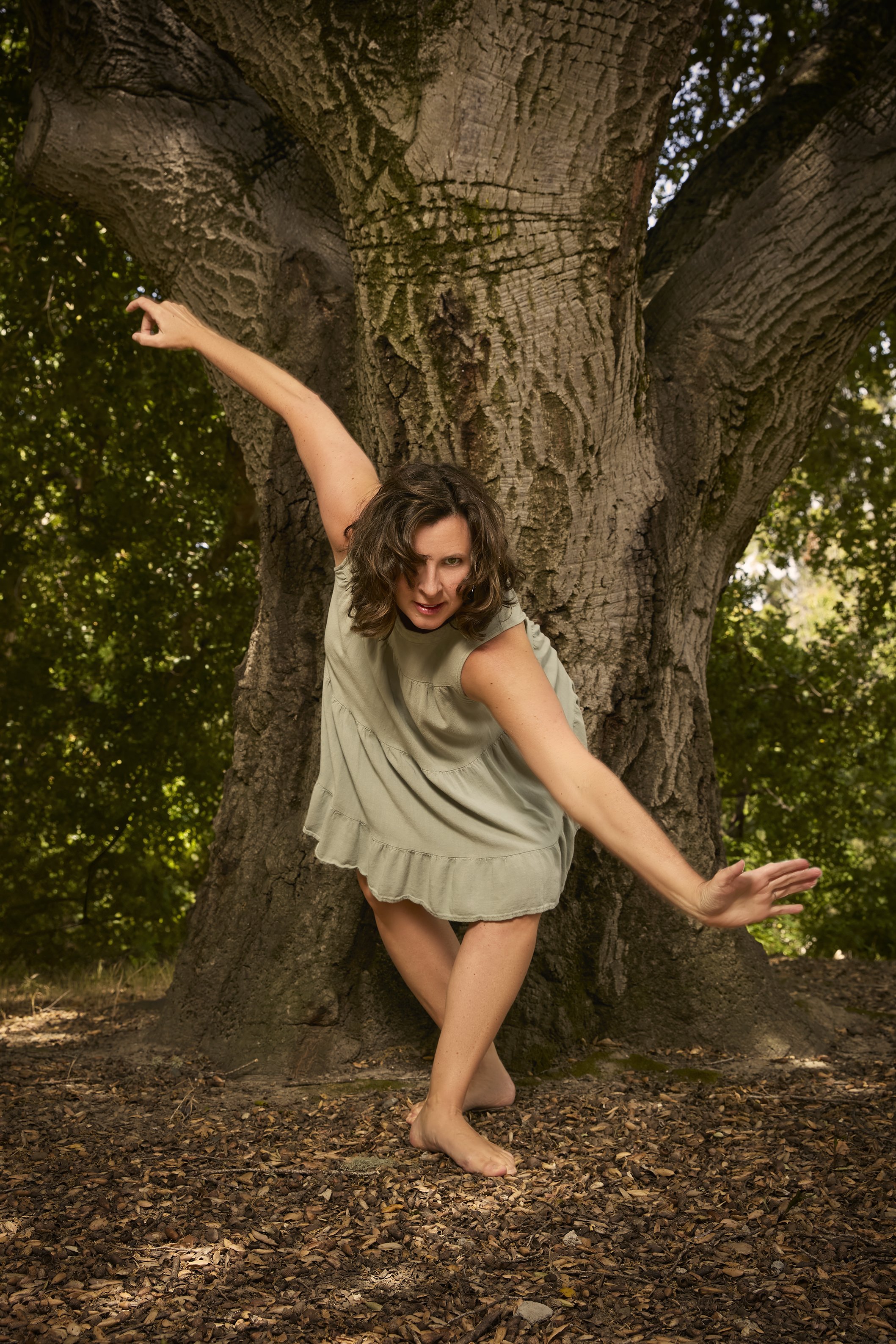 A woman with wavy brown hair wearing a light green dress, barefoot, posing playfully in front of a large tree with dense green foliage in the background.