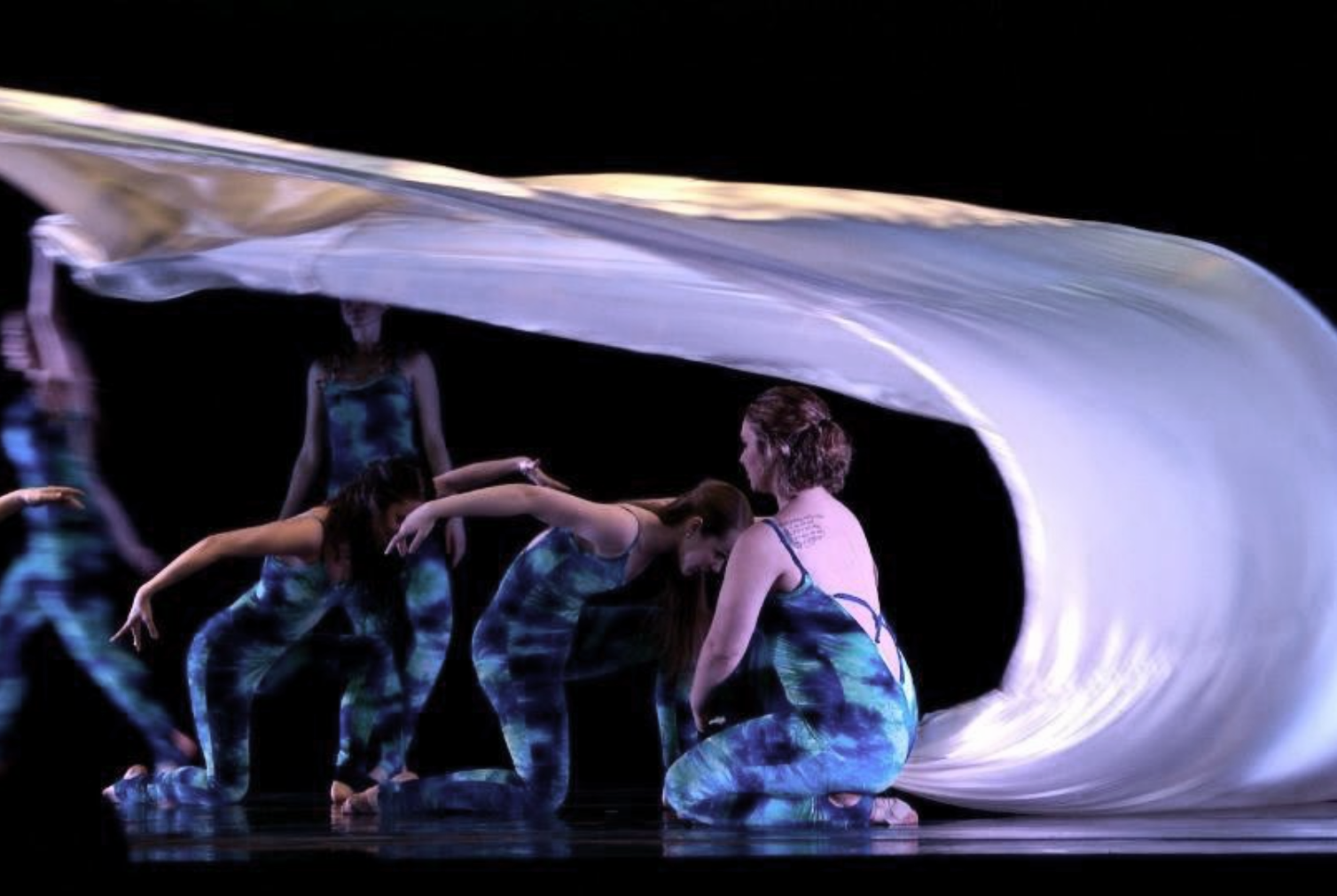 A group of dancers in colorful, patterned costumes performing on stage with flowing fabric overhead, creating a dynamic and artistic scene.