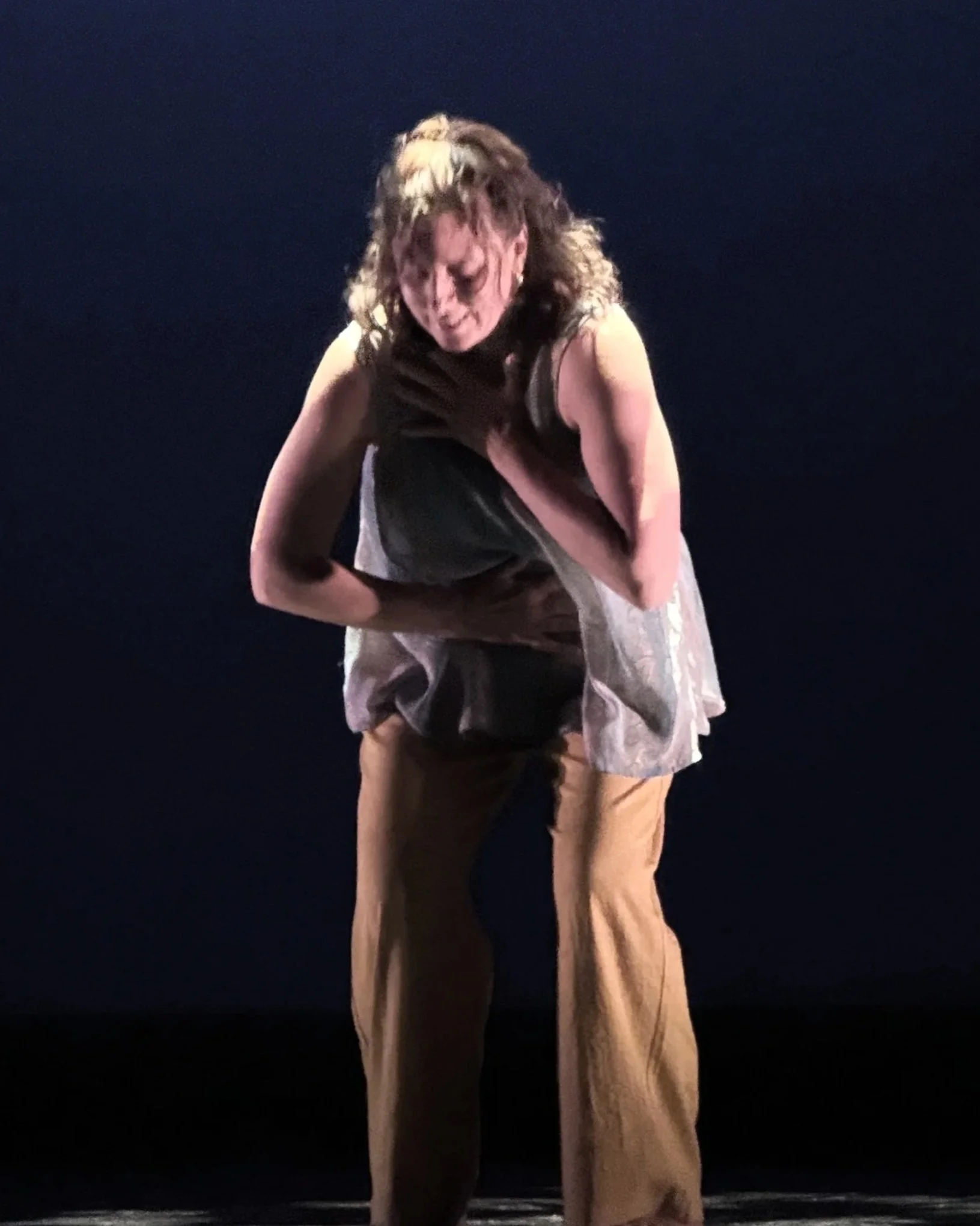 A woman with curly hair wearing a sleeveless top and tan pants is bent over, clutching her chest with a pained expression against a dark background.