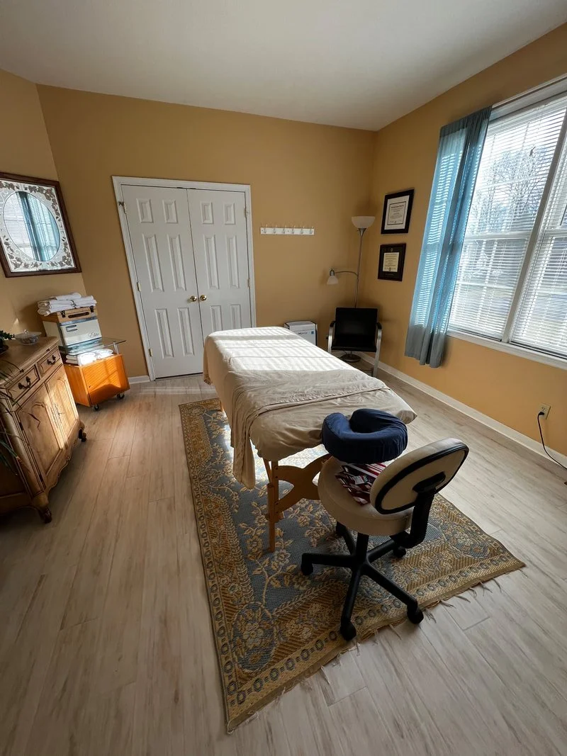 A massage or treatment room with a massage table covered in cream-colored sheet, an office chair with a stack of folded towels, a wooden cabinet, and a wall with framed certificates or diplomas, a floor lamp, a rocking chair, and a large window with blue curtains letting in natural light.