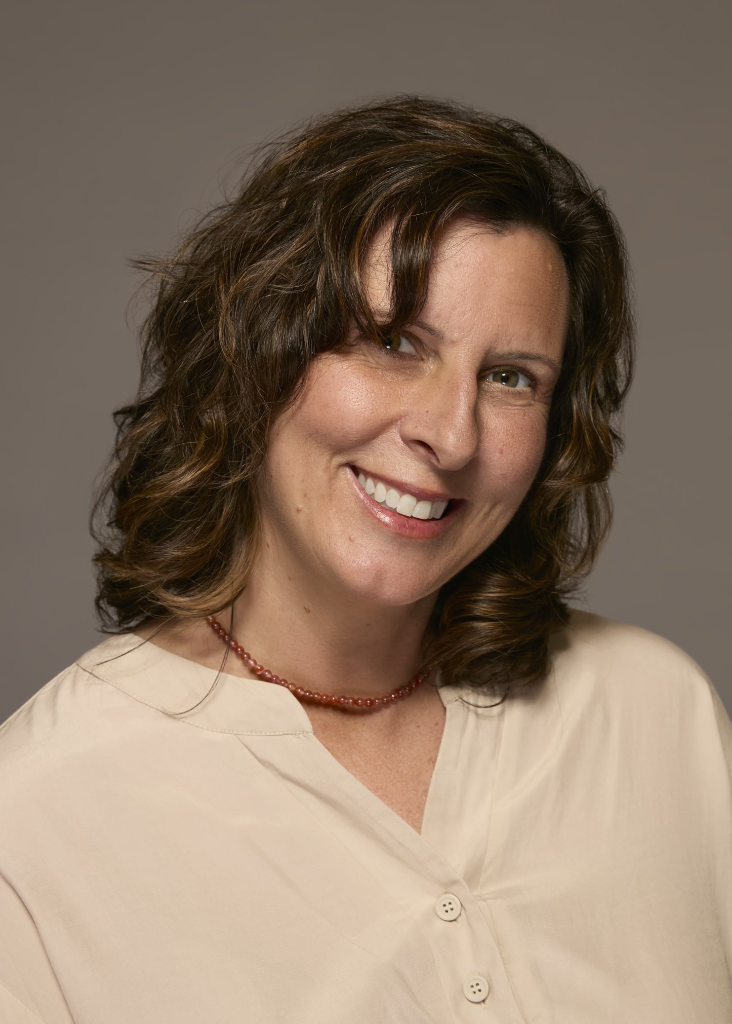 A woman with shoulder-length brown hair, smiling, wearing a cream blouse and a red beaded necklace, against a gray background.