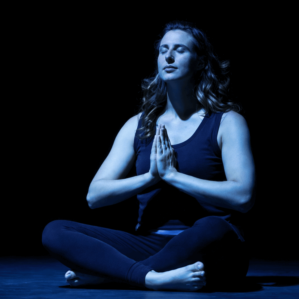 A woman practicing yoga or meditation, seated in a cross-legged position, with eyes closed and hands pressed together in prayer gesture, illuminated in blue lighting.