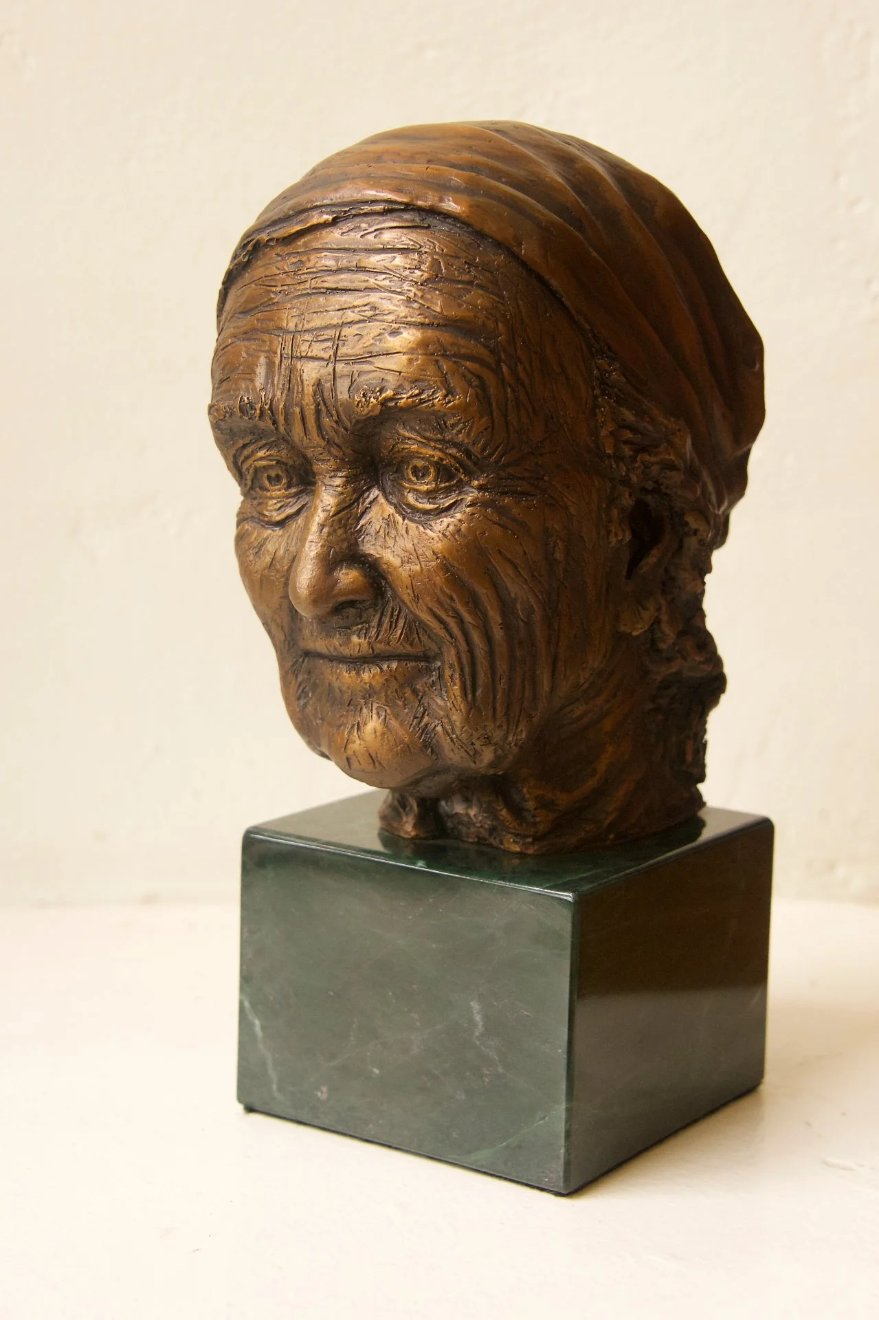 Wooden bust sculpture of an elderly woman with a headscarf on a dark marble pedestal.