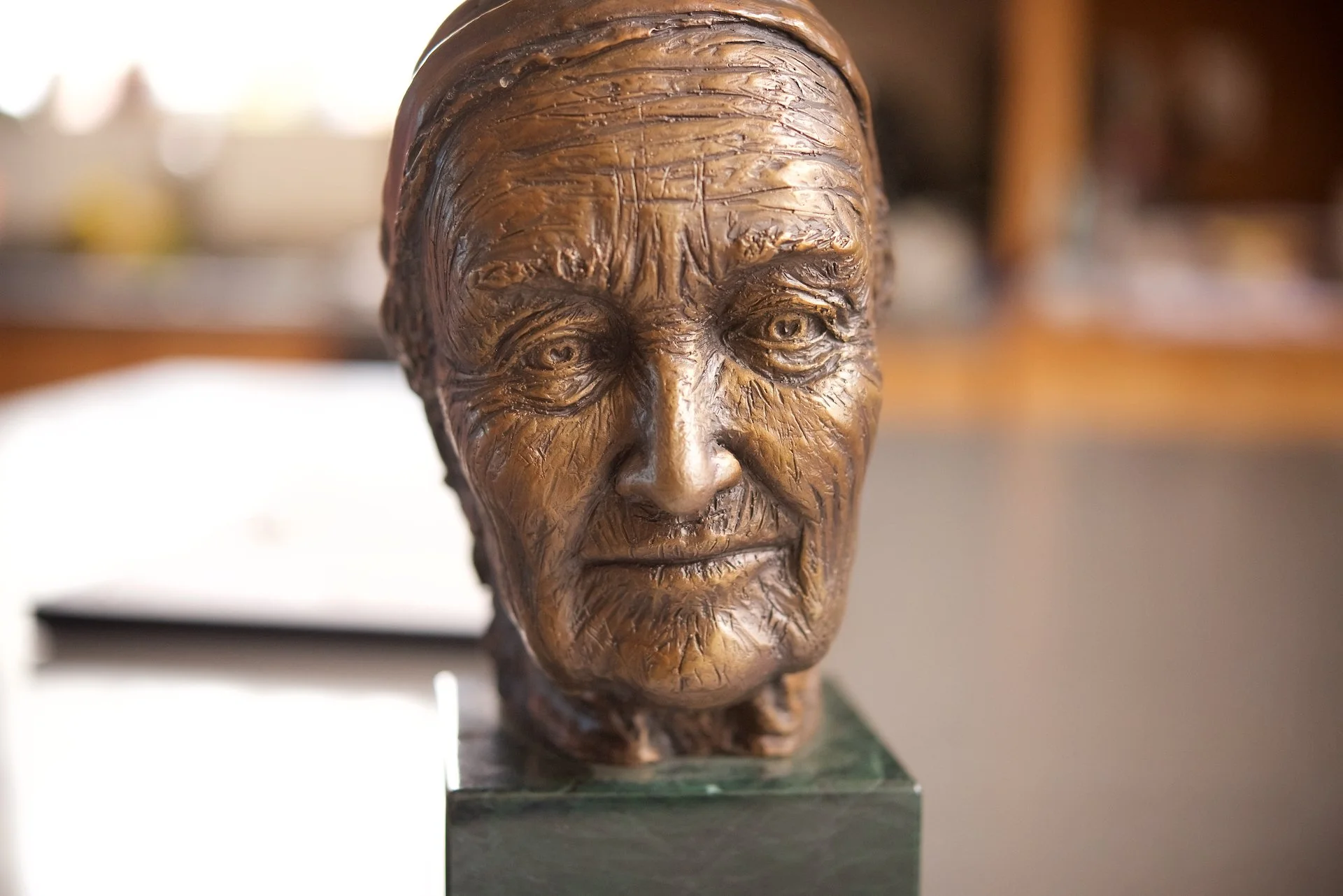 Bronze bust sculpture of an elderly woman with detailed wrinkles and expression, placed on a green marble base in an indoor setting.