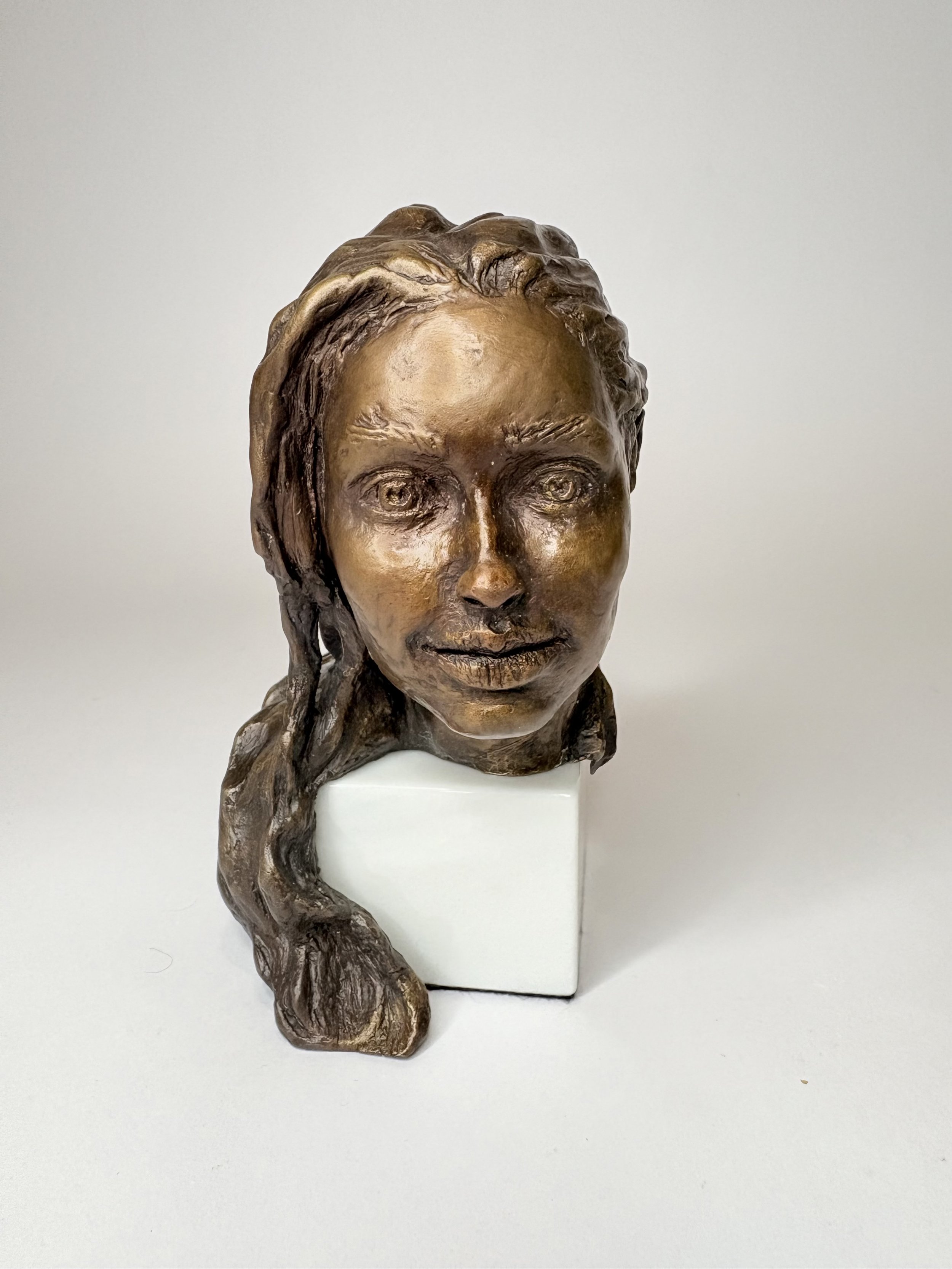 Bronze sculpture of a woman's face with long hair, mounted on a white pedestal.