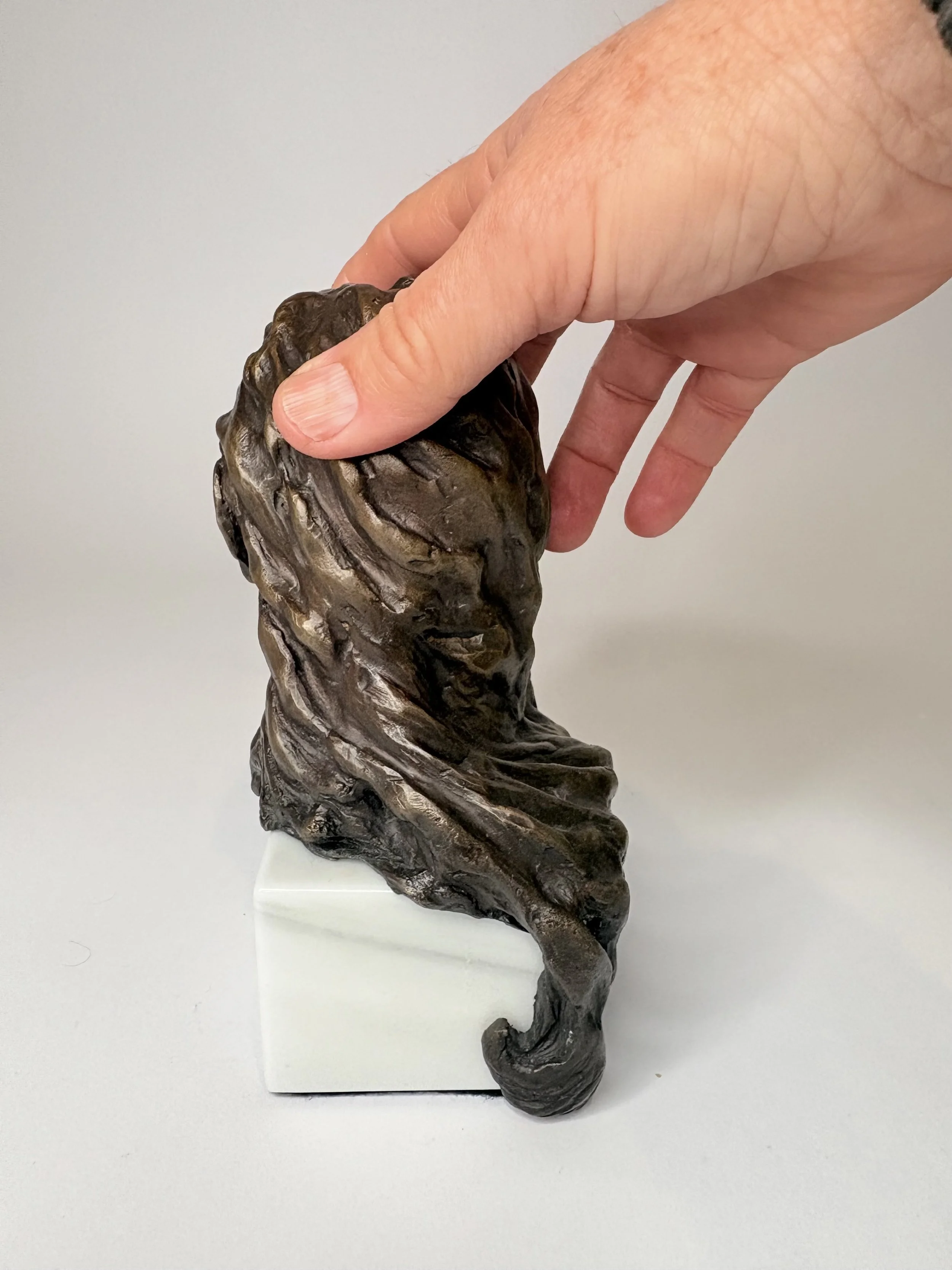 Hand holding a small sculpture of a person's head with long hair on a white base.