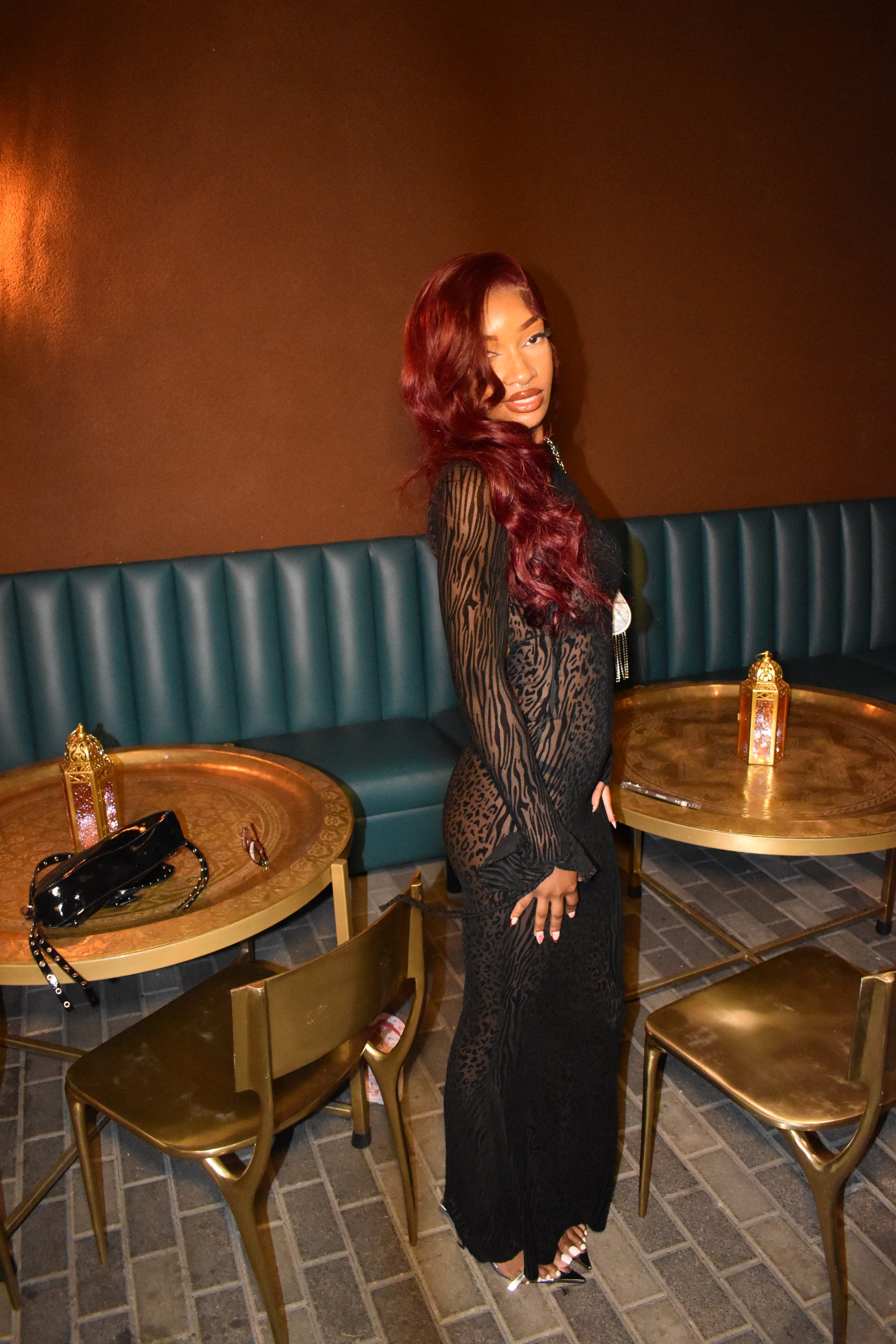 A woman with long reddish-brown hair standing indoors near a teal leather banquette, with gold tables and chairs around her, wearing a sheer black dress with animal print details.