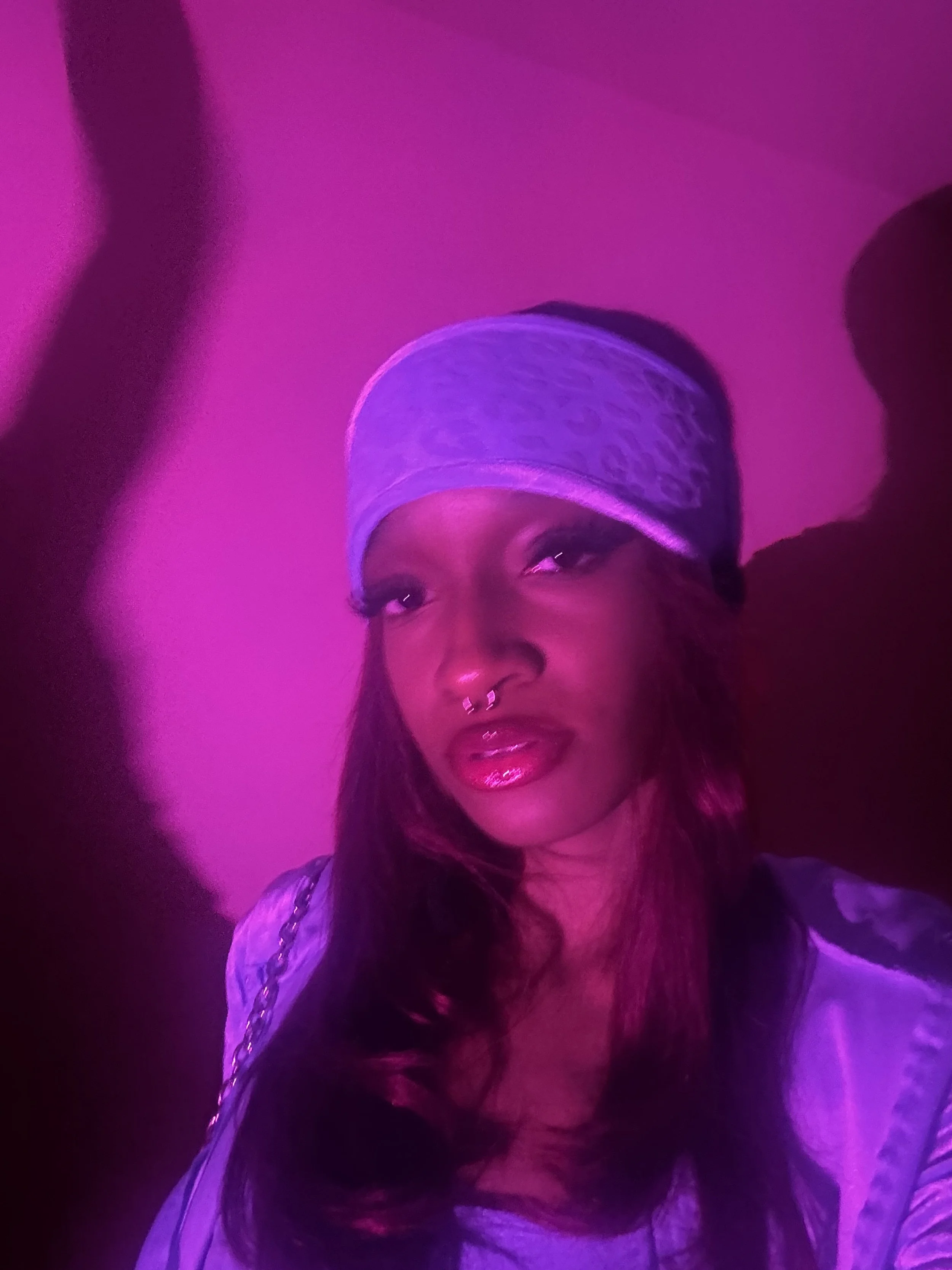 A woman with long hair, wearing a light purple bandana and a satin jacket, is illuminated by purple-pink lighting and looking into the camera.