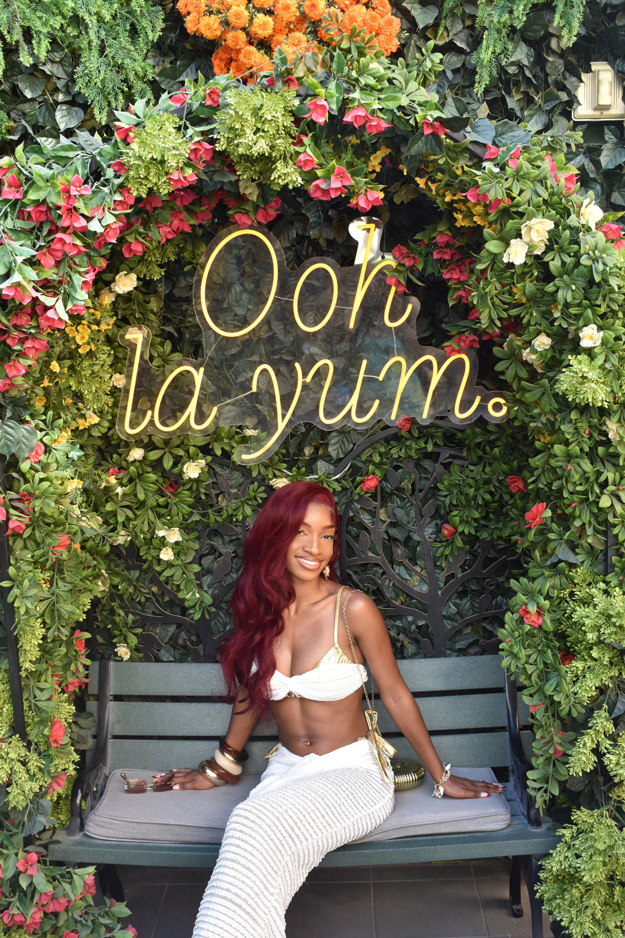 A woman with long red hair smiling while sitting on a gray bench in front of a floral backdrop. The backdrop features a variety of green foliage, colorful flowers, and a neon sign that reads 'Ooh la yum.'