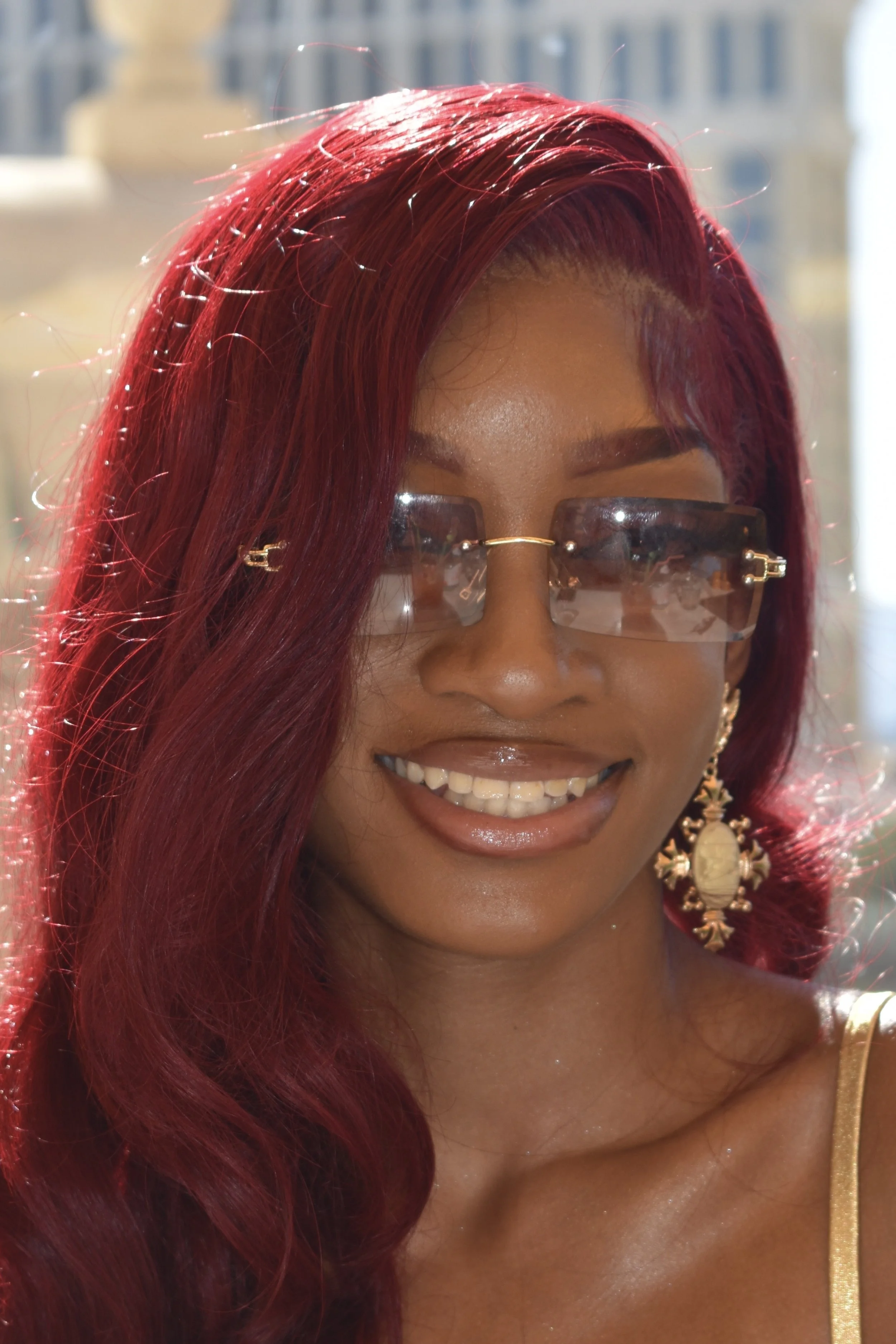 A woman with long red hair styled in waves, wearing fashionable sunglasses, and large ornate earrings, smiling outdoors in bright sunlight.