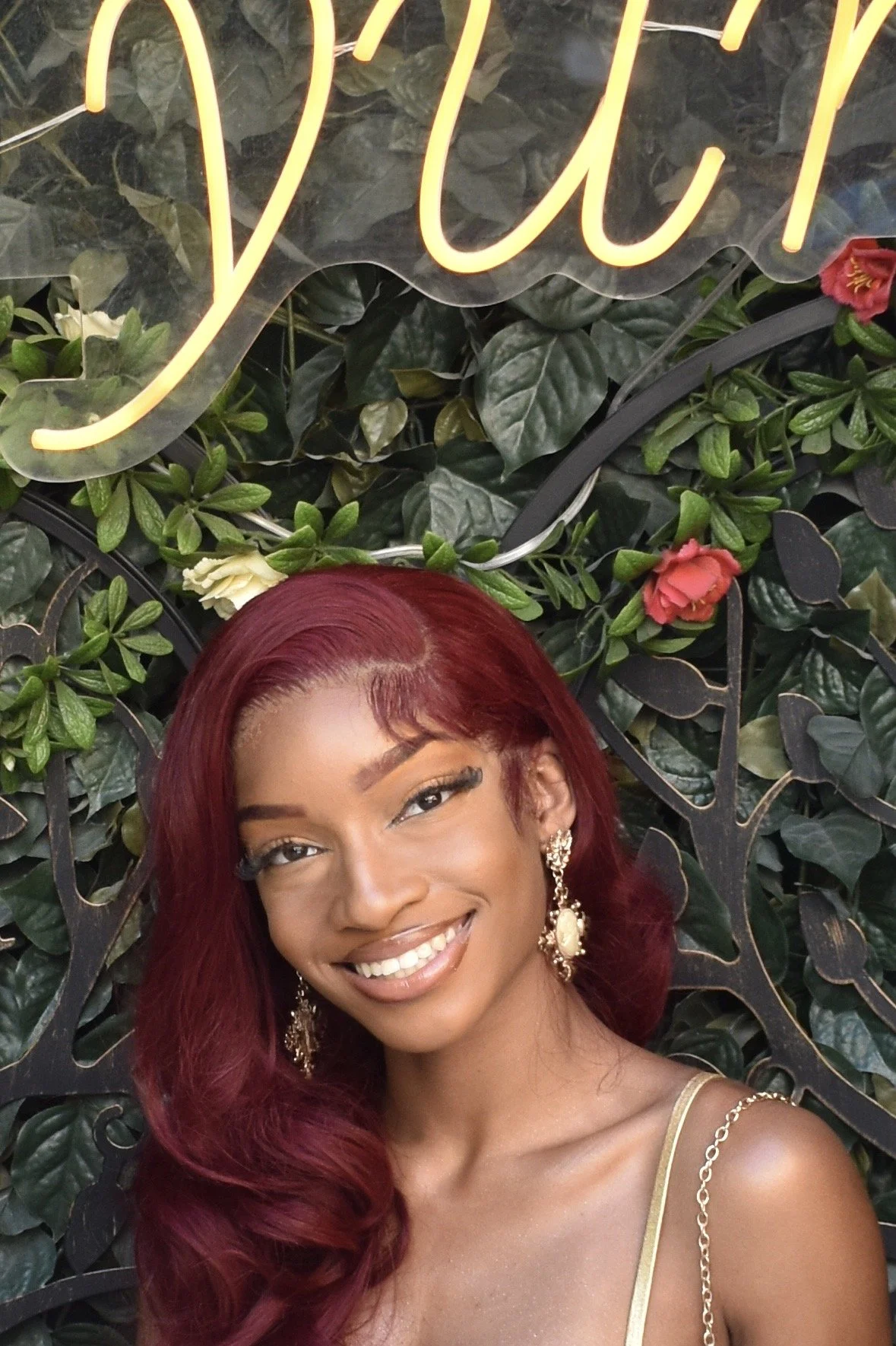 A smiling woman with long burgundy hair, wearing gold earrings and a gold strap dress, standing in front of a green leafy background and a neon sign.