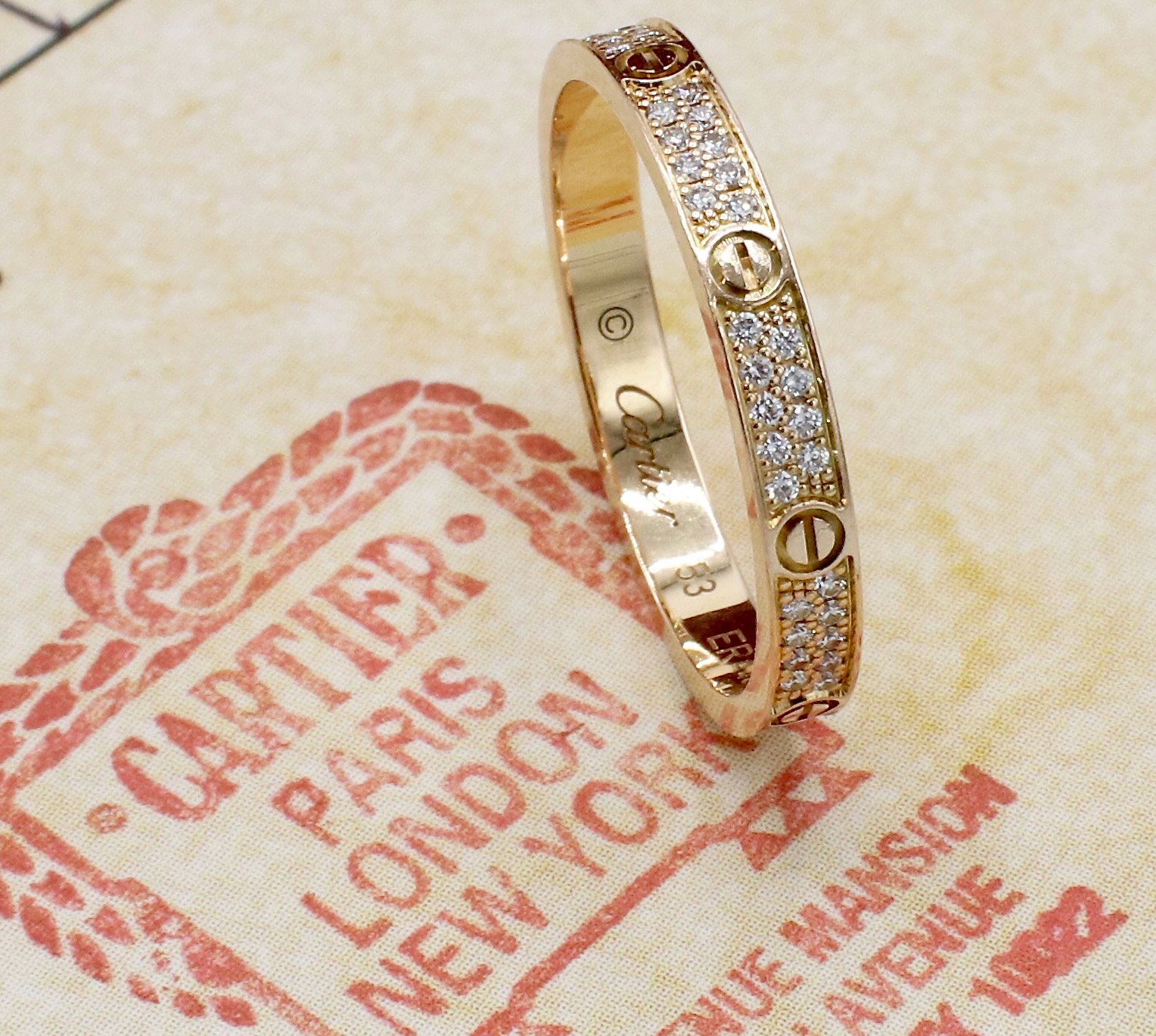 Gold Cartier Love ring with embedded small diamonds, resting on a fabric with Cartier branding and red decorative elements.