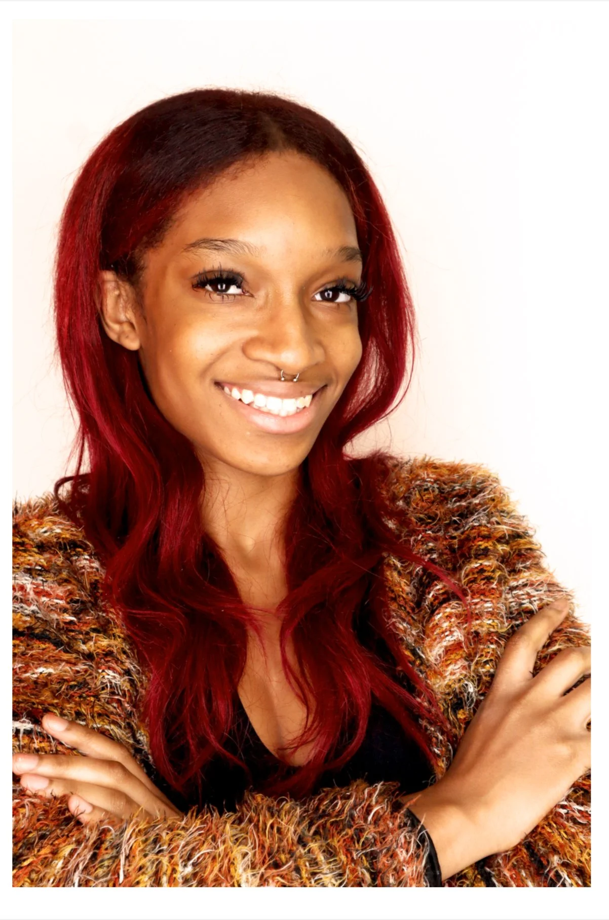A young woman with long red hair, a nose septum piercing, and a warm brown complexion, smiling with crossed arms, wearing a multicolored textured sweater against a plain white background.
