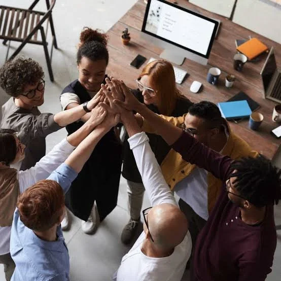 Group of diverse coworkers standing in a circle, high-fiving each other in a modern office with desks, computer monitors, and office supplies.