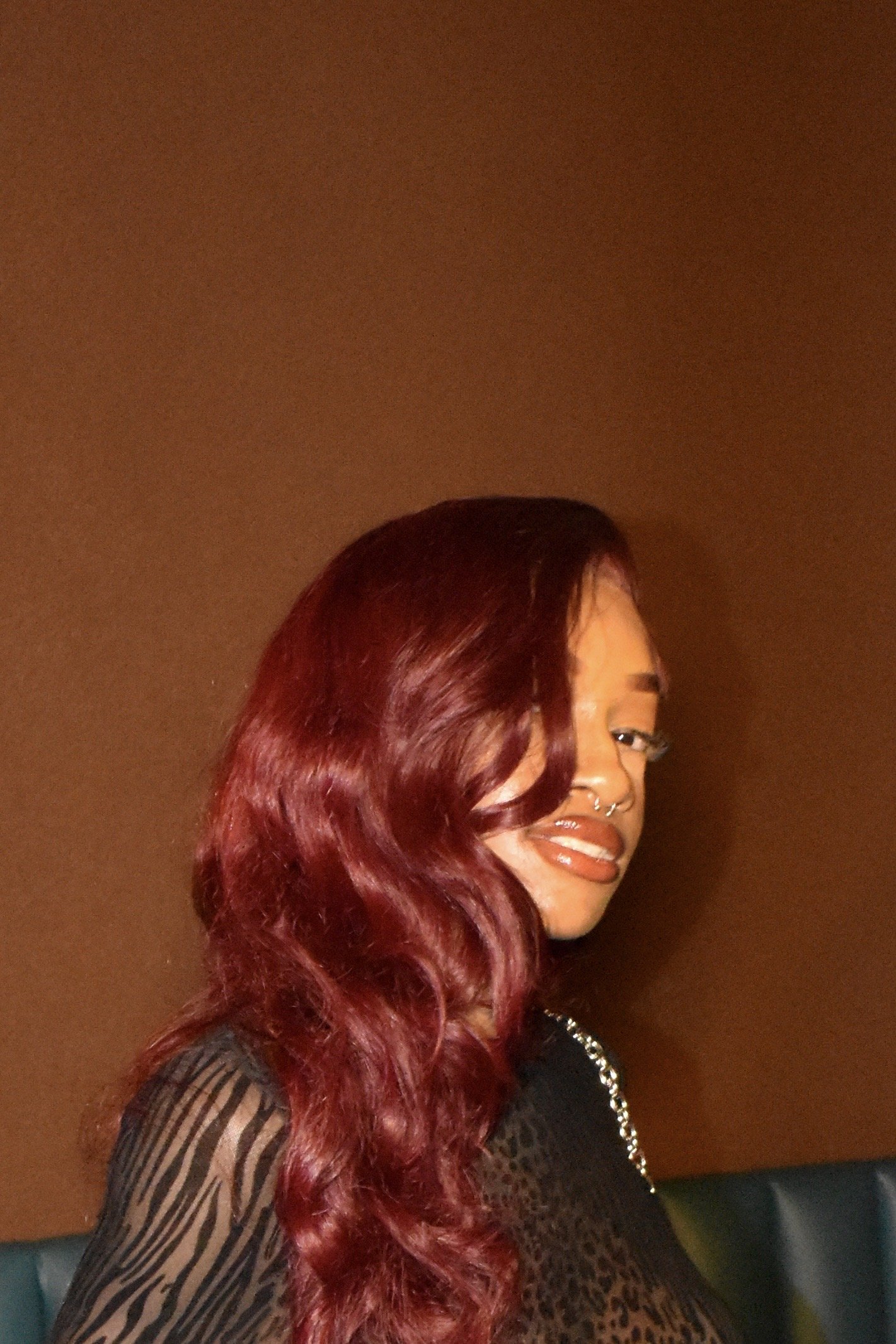 A woman with long wavy red hair, wearing a mesh top with animal print pattern, and sporting a septum piercing, lipstick, and eye makeup pose against a brown wall.