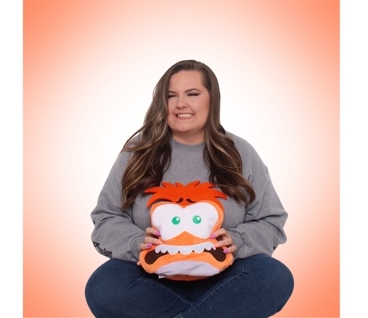 A woman with long brown hair, wearing a gray sweatshirt and jeans, sitting cross-legged, holding a plush toy of a cartoonish, orange, angry face with big eyes and a furrowed brow, against an orange background.