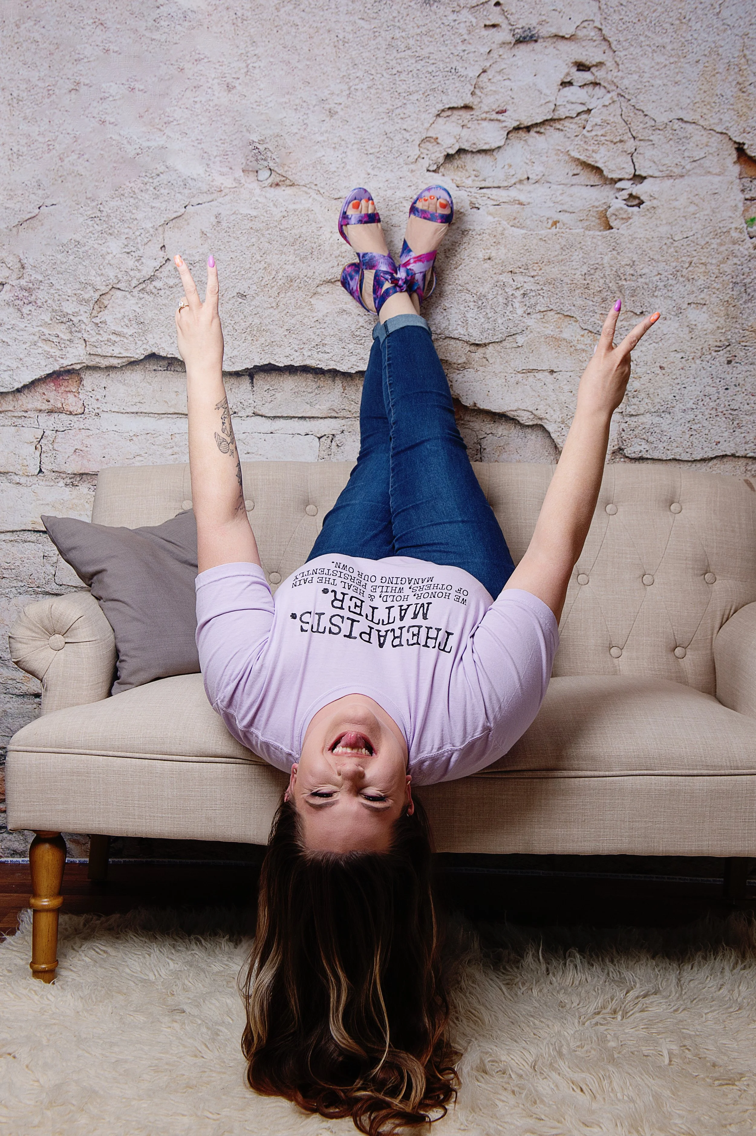 A woman with long hair sticking her tongue out, lying upside down on a sofa with her legs raised against a textured wall, making peace signs with her hands, wearing a white T-shirt with black text, blue jeans, and colorful high-heeled sandals.