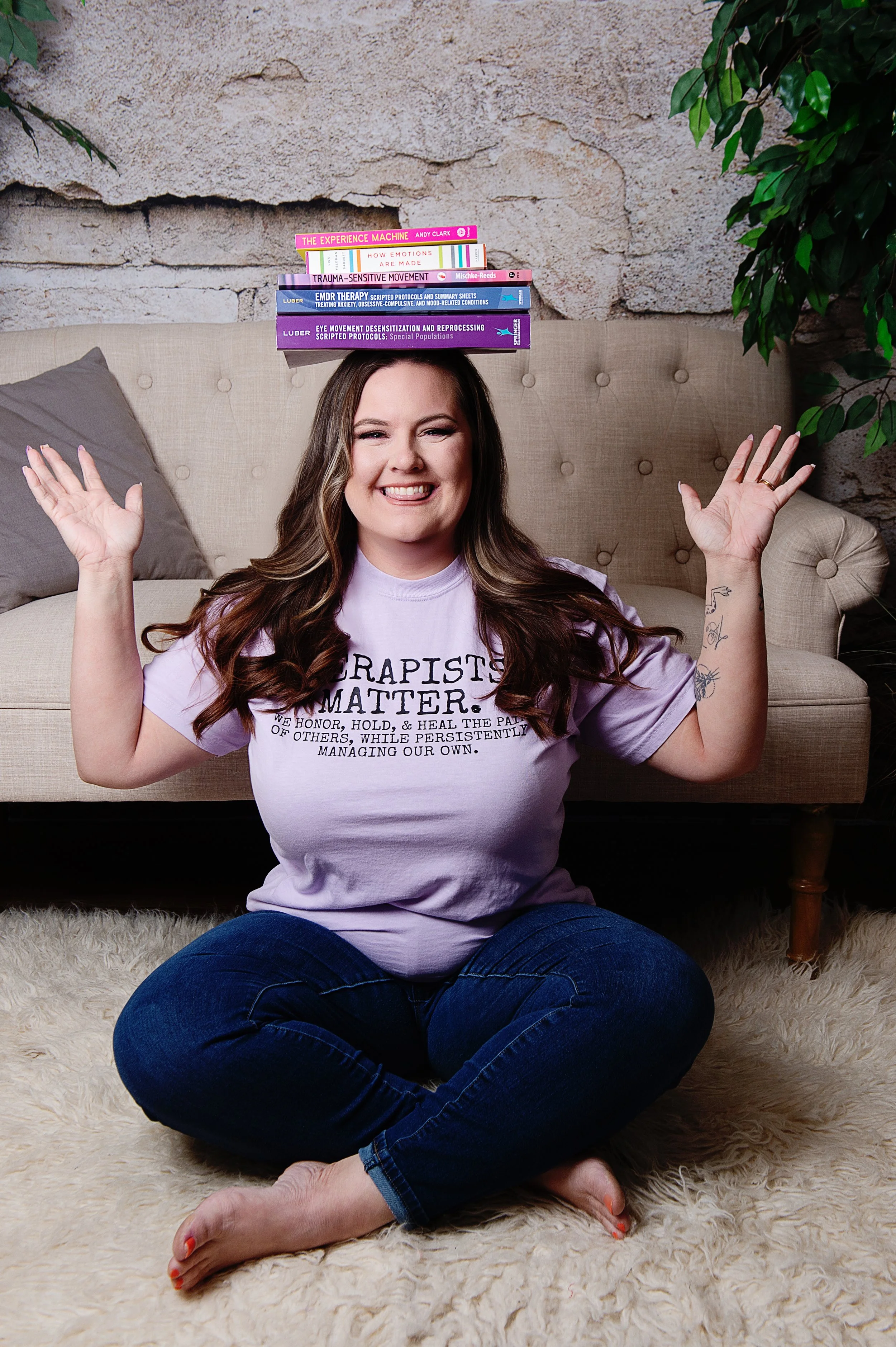 A woman with long brown hair sitting cross-legged on a fluffy cream rug in front of a beige couch. She is smiling and has her hands raised. She wears a lavender T-shirt with the words 'Grapists Matter' and a list of words underneath, with a stack of books balanced on her head. The background includes a stone wall, a gray pillow on the couch, and a green plant on the right.