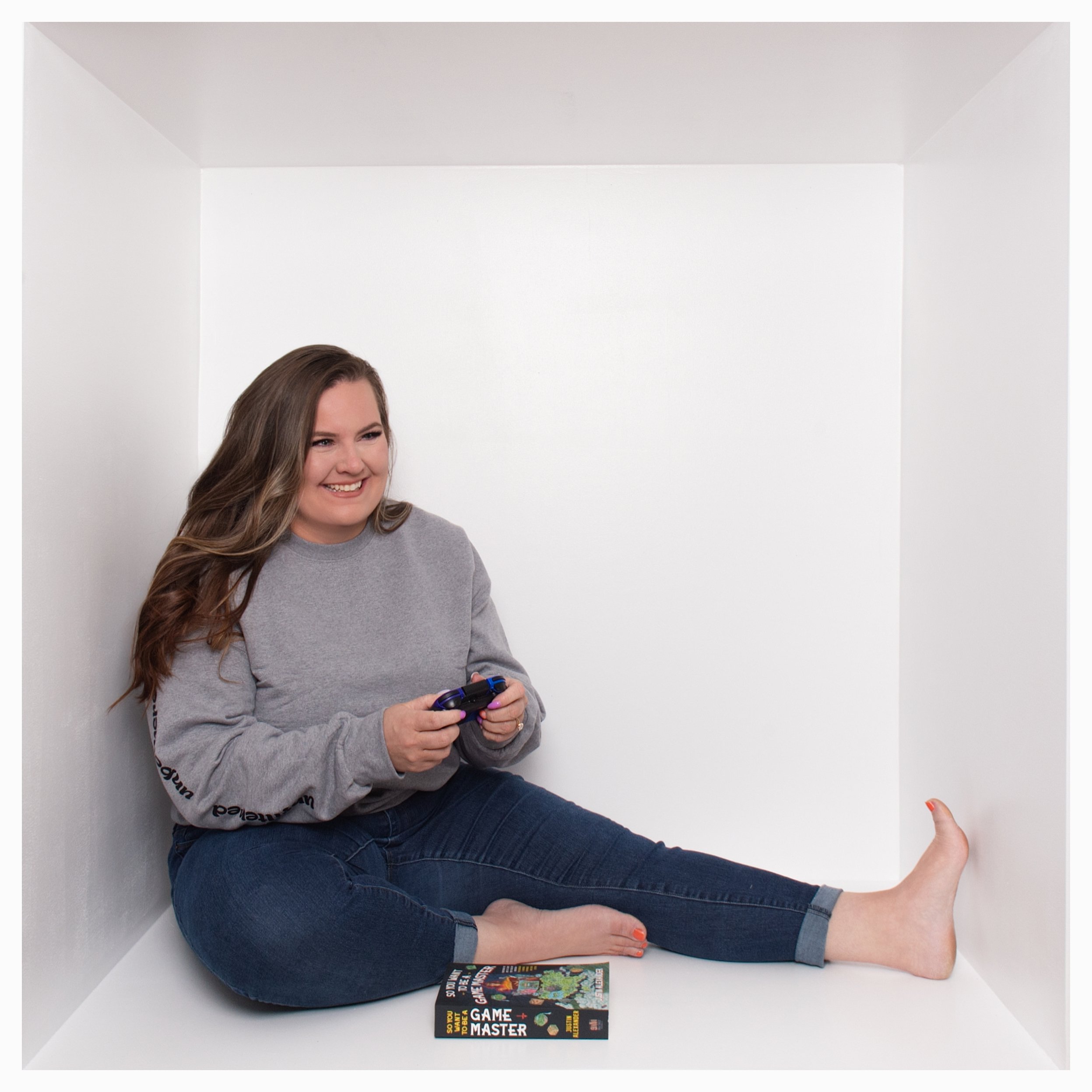 A woman sitting in a white box, smiling and holding a video game controller, with a book titled 'Game Master' on the floor beside her.