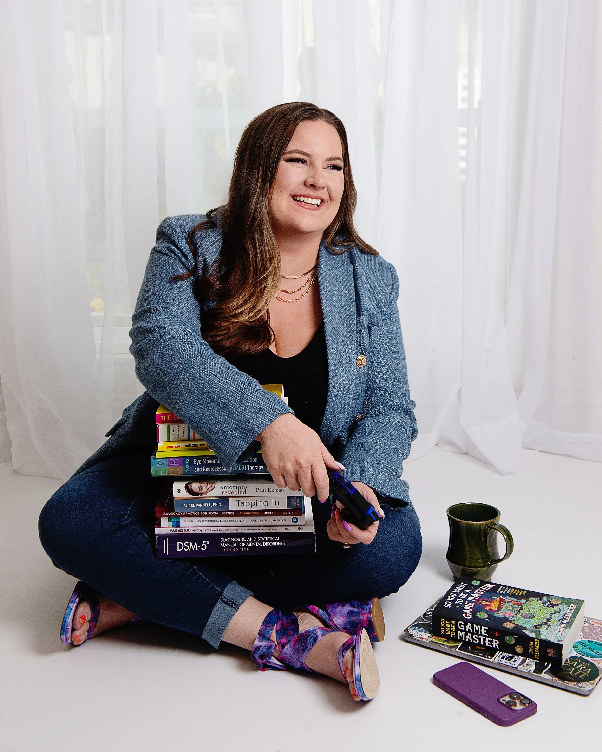 A woman with long brown hair sitting cross-legged on the floor, smiling, wearing a blue blazer over a black top, surrounded by books, a mug, and a phone.