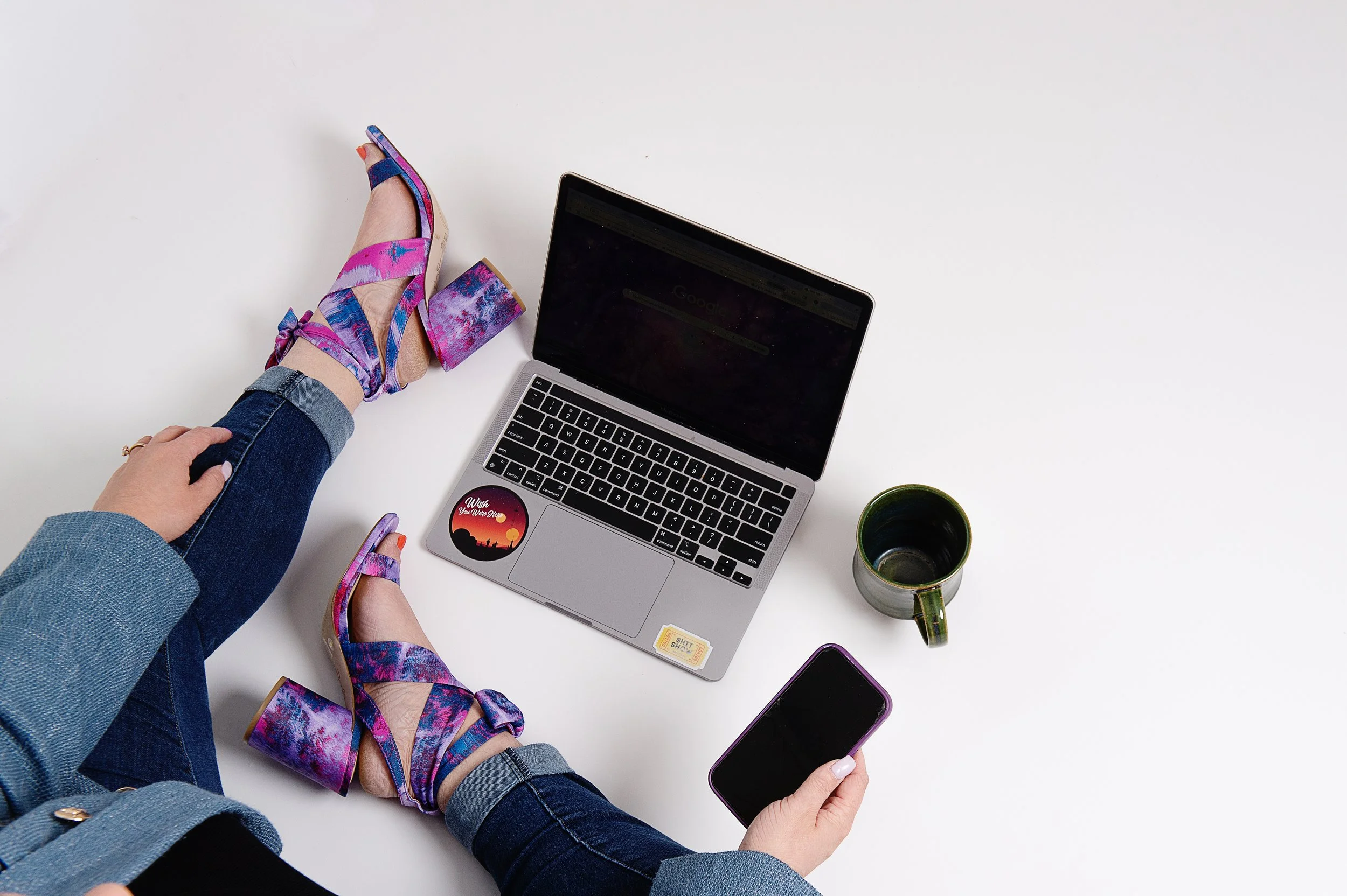 Person wearing colorful high-heeled sandals, blue jeans, and a denim jacket, holding a smartphone, with a laptop, a green coffee mug, and a purple phone on a white surface.