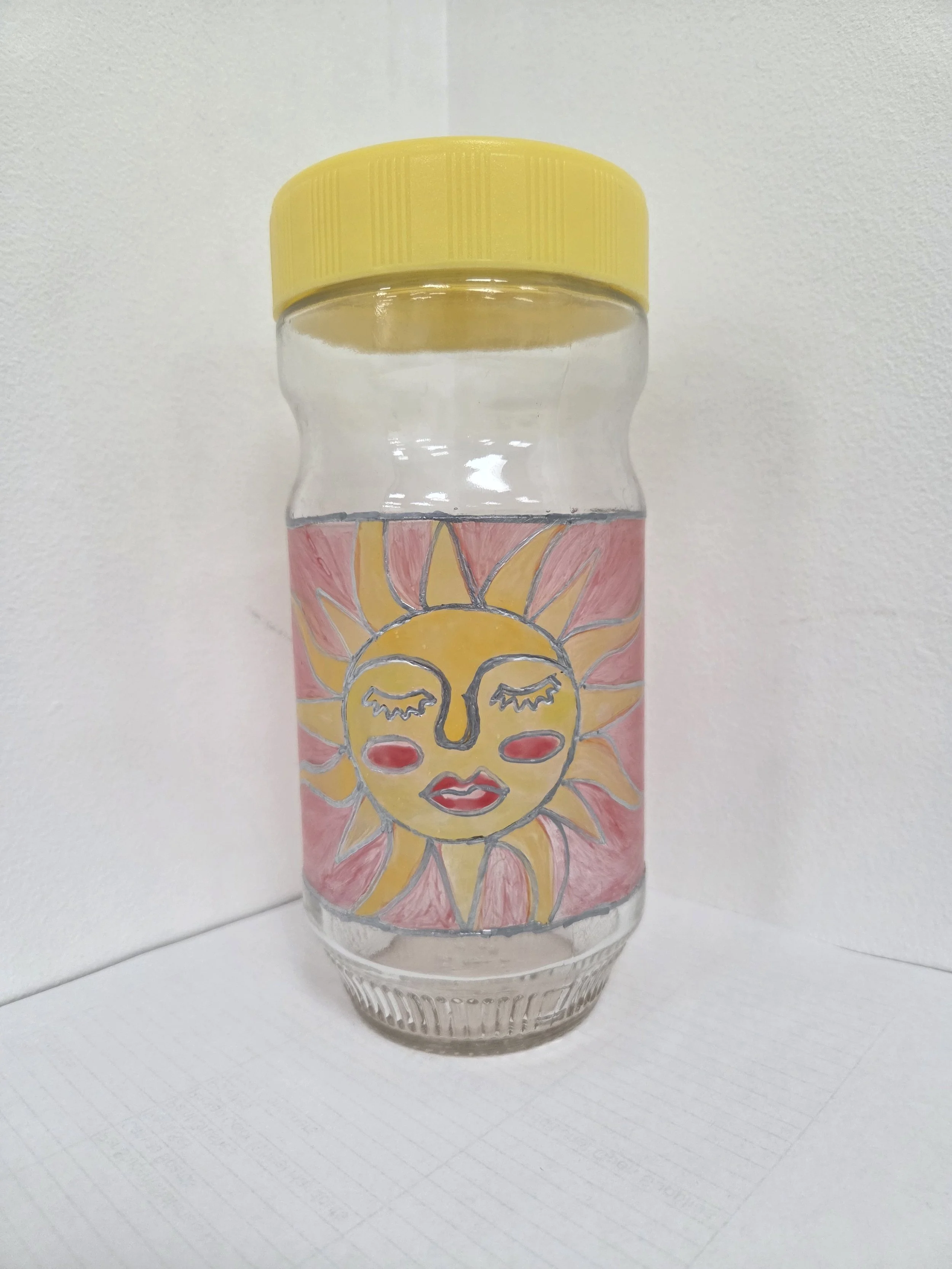 A clear glass jar with a yellow cap and a colorful glass painting of a sun with a face.