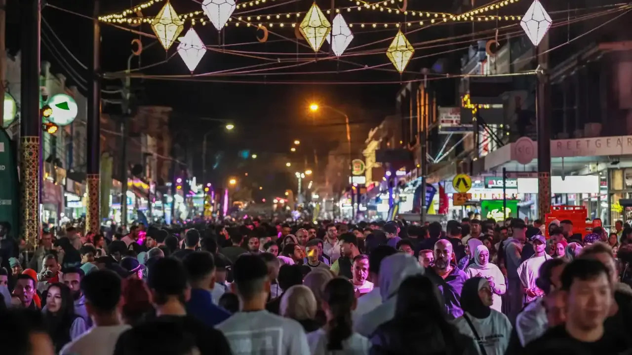 A crowded city street at night with street lights, colorful neon signs, and hanging lanterns or decorations overhead during Lakemba Nights during Ramadan.
