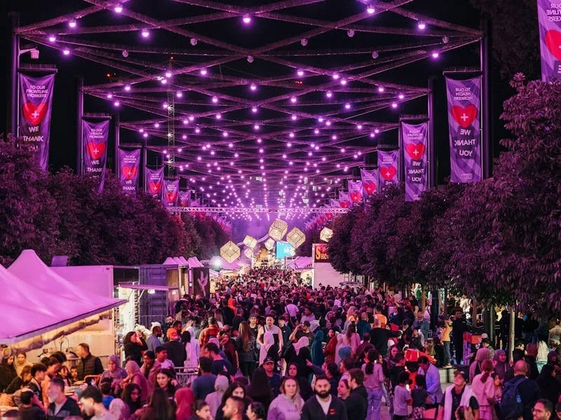 A large outdoor festival at night with purple lights hanging overhead, crowd of people, market stalls, and banners with red hearts and white text.
