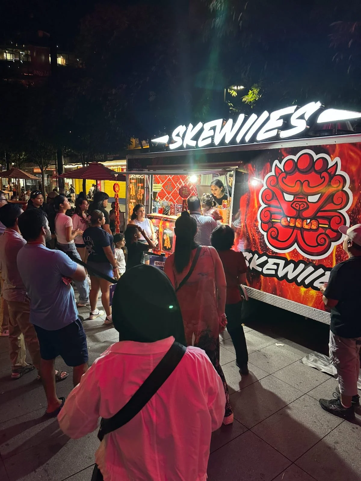 A nighttime scene of a food truck named 'Skewies' serving customers outdoors. A line of people waits to order, with some standing and others facing the truck window, which is illuminated. The truck has a bold red and black design with a fierce lion graphic. Several red umbrellas provide shade to nearby groups of people.