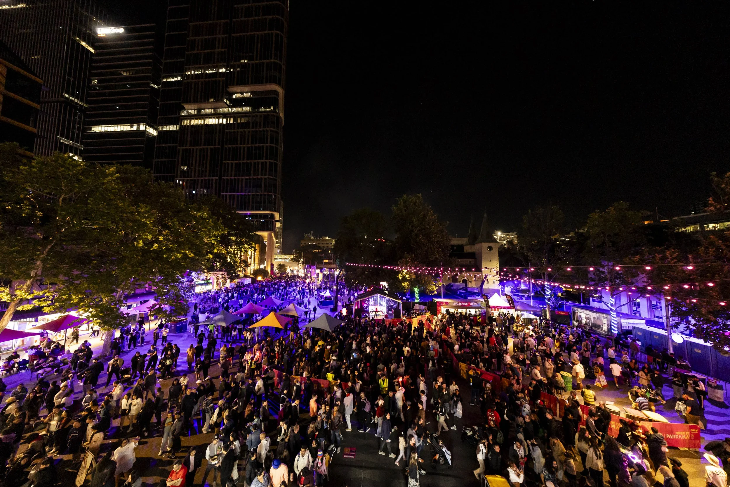 Nighttime street festival Parramatta Lanes with a large crowd, colorful lights, and tents in an urban area with tall buildings and trees.
