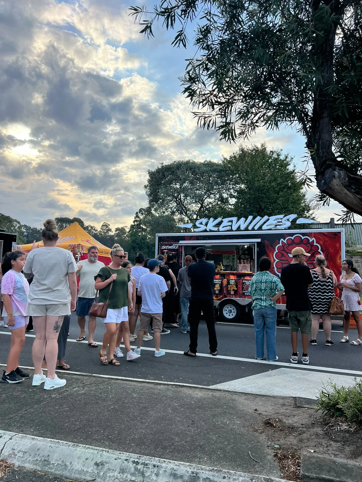 People waiting in line at a food truck named 'Skewiies' at an outdoor event during late afternoon or early evening, with trees and a partly cloudy sky in the background. This is for Campbelltown NYE at Koshigaya Park