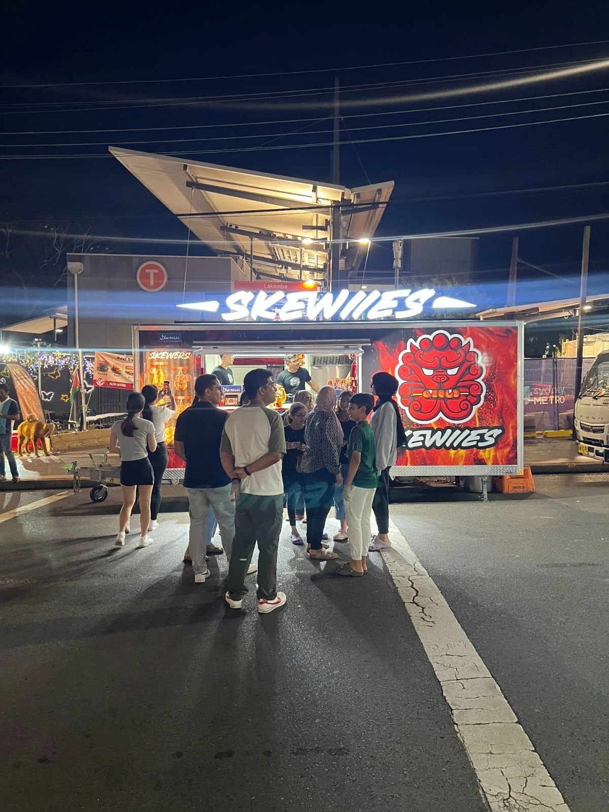 Night scene of a busy outdoor food stand named 'Skewiies' with a group of people waiting in line and socializing, illuminated signage, and a small food vending area.