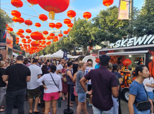 Crowd of people at an outdoor event with string lights and red lanterns hanging overhead, with Skewiies food truck and trees in the background.