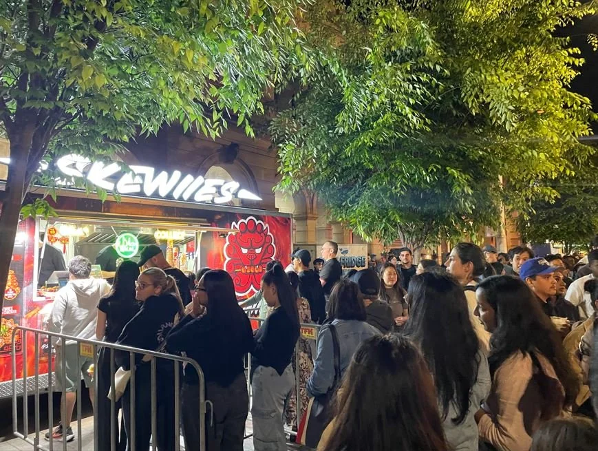 Crowd of people standing in line at Skewiies food truck at night under green trees, with the truck illuminated and a fire-themed logo during Parramatta Lanes.