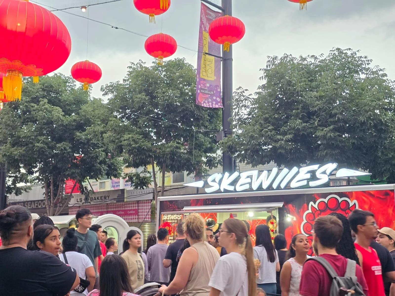 Crowd of people gathering at an outdoor event with red Chinese lanterns hanging overhead and Skewiies food truck with a neon sign that reads 'SKEWIIES'.