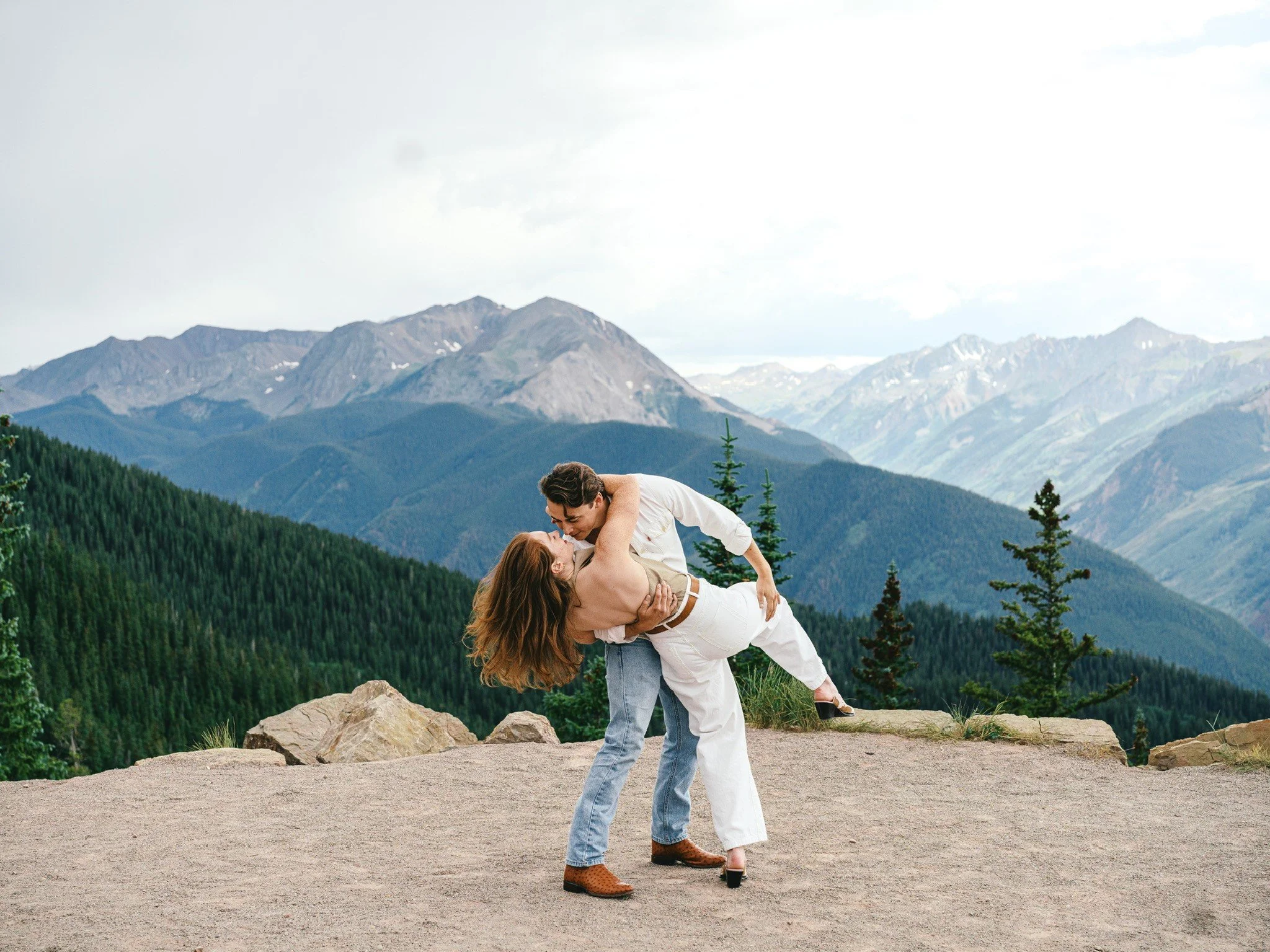 A couple dancing outdoors on a rocky surface with a scenic mountain range and forest in the background.