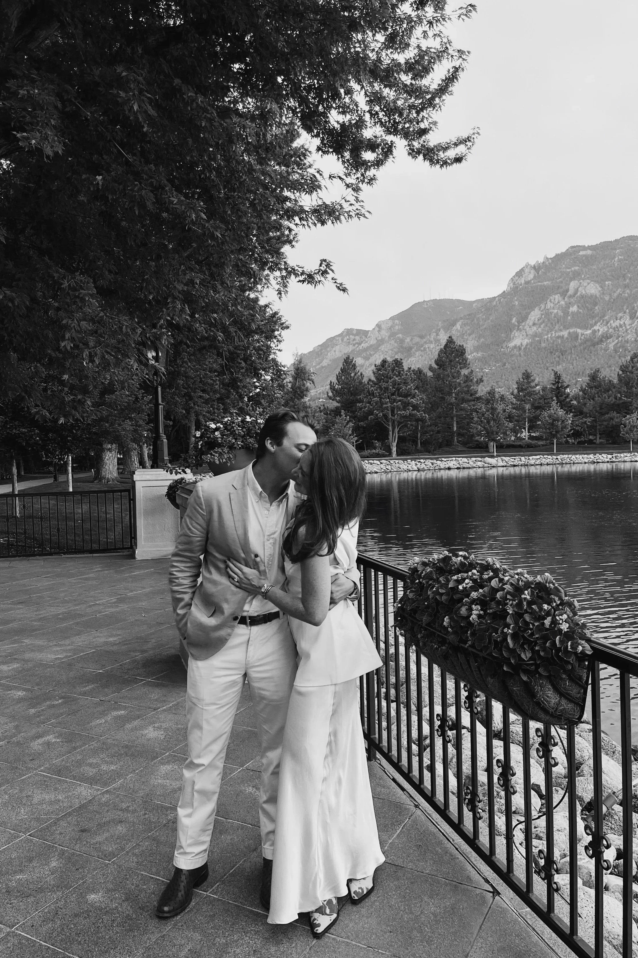 A couple sharing a kiss by a lake at sunset, surrounded by trees and mountains, in black and white.