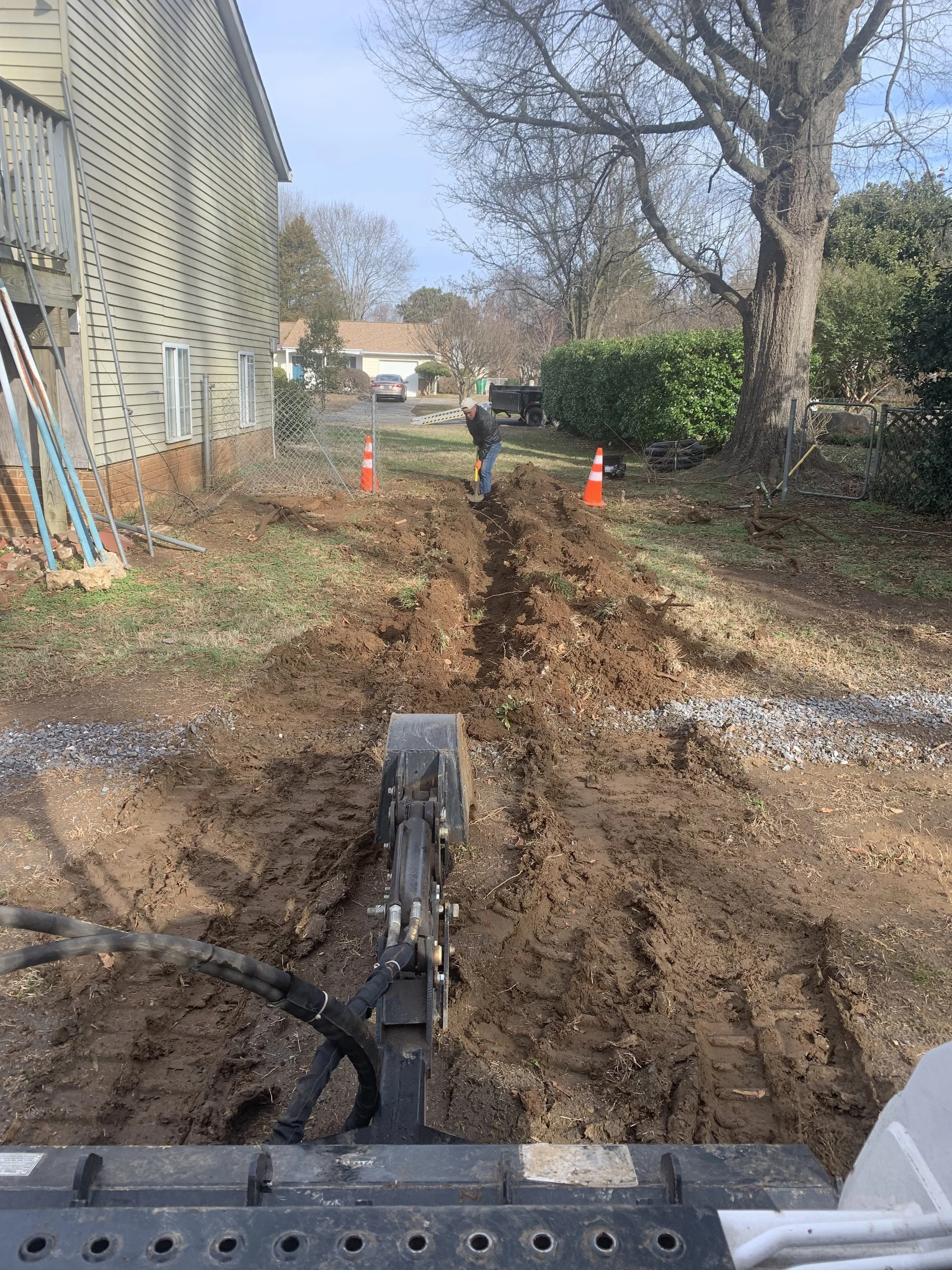 A construction worker operates a small excavator digging a trench in a residential yard, with orange traffic cones and a man working in the background.