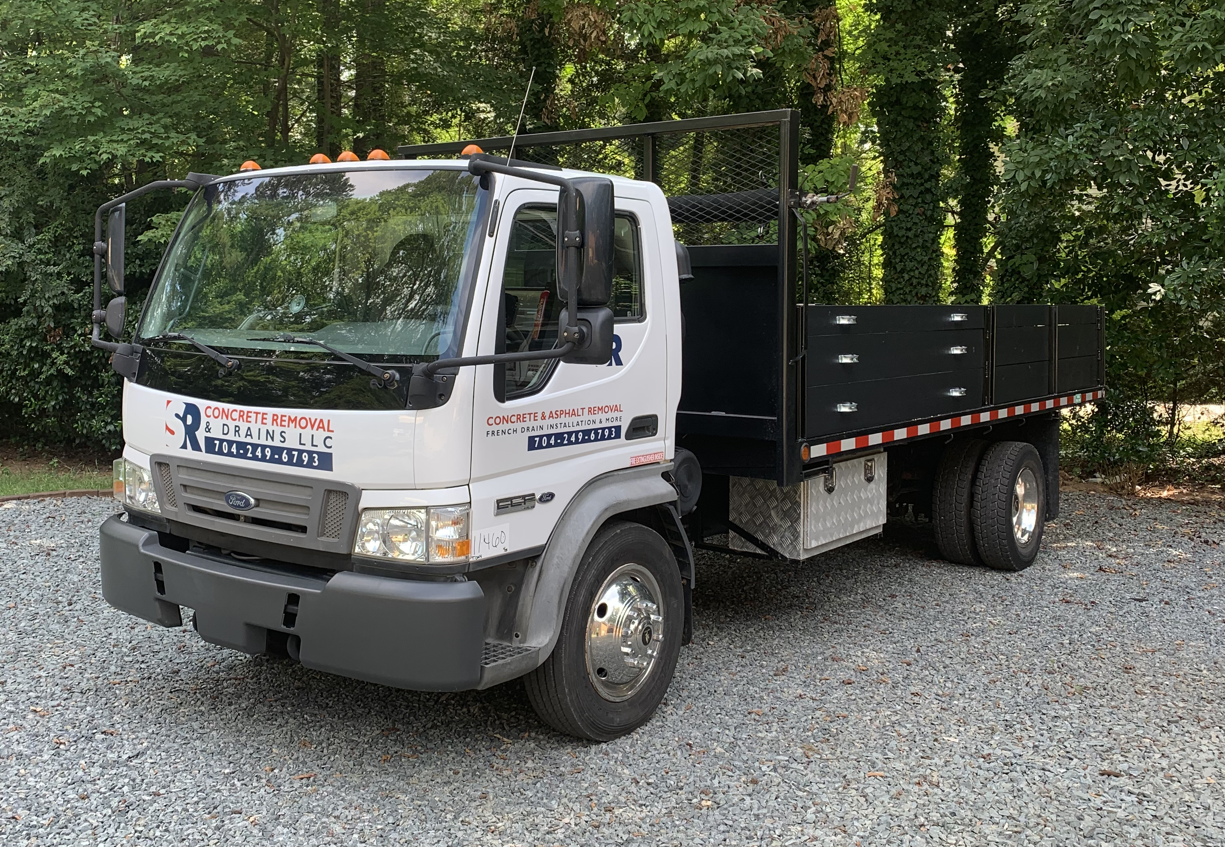 A white flatbed truck parked on a gravel surface surrounded by green trees, with signage indicating it is for concrete removal and drainage services.