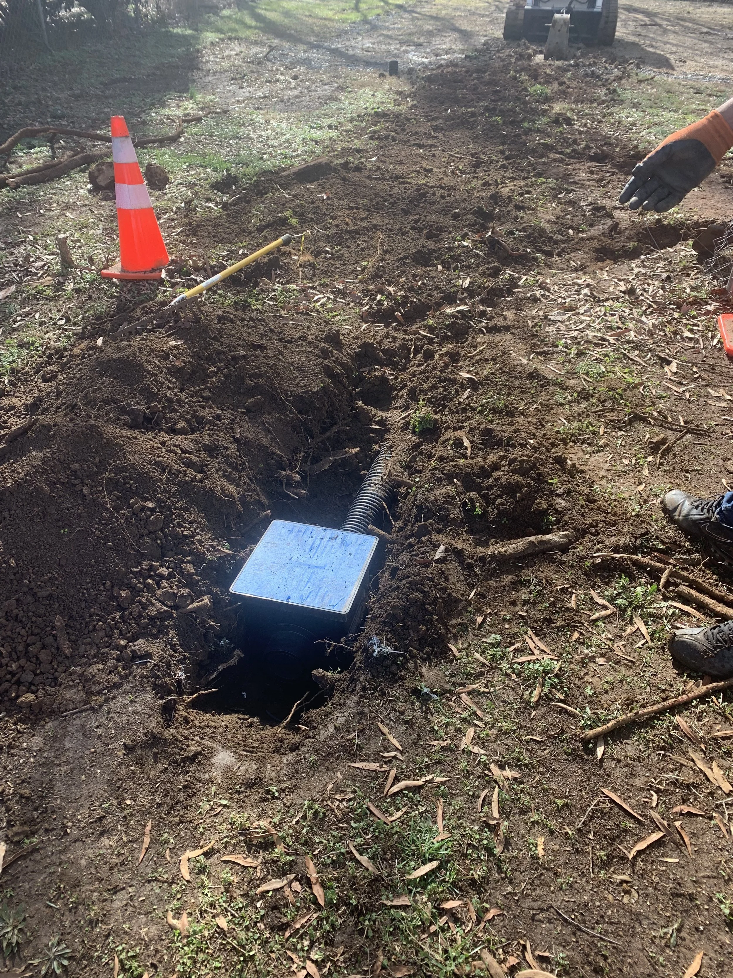 A utility worker's hand, wearing a glove, is near a recently dug hole with an underground cable or conduit. An orange safety cone, a yellow measuring tool, and a small excavator are visible nearby on the dirt ground.