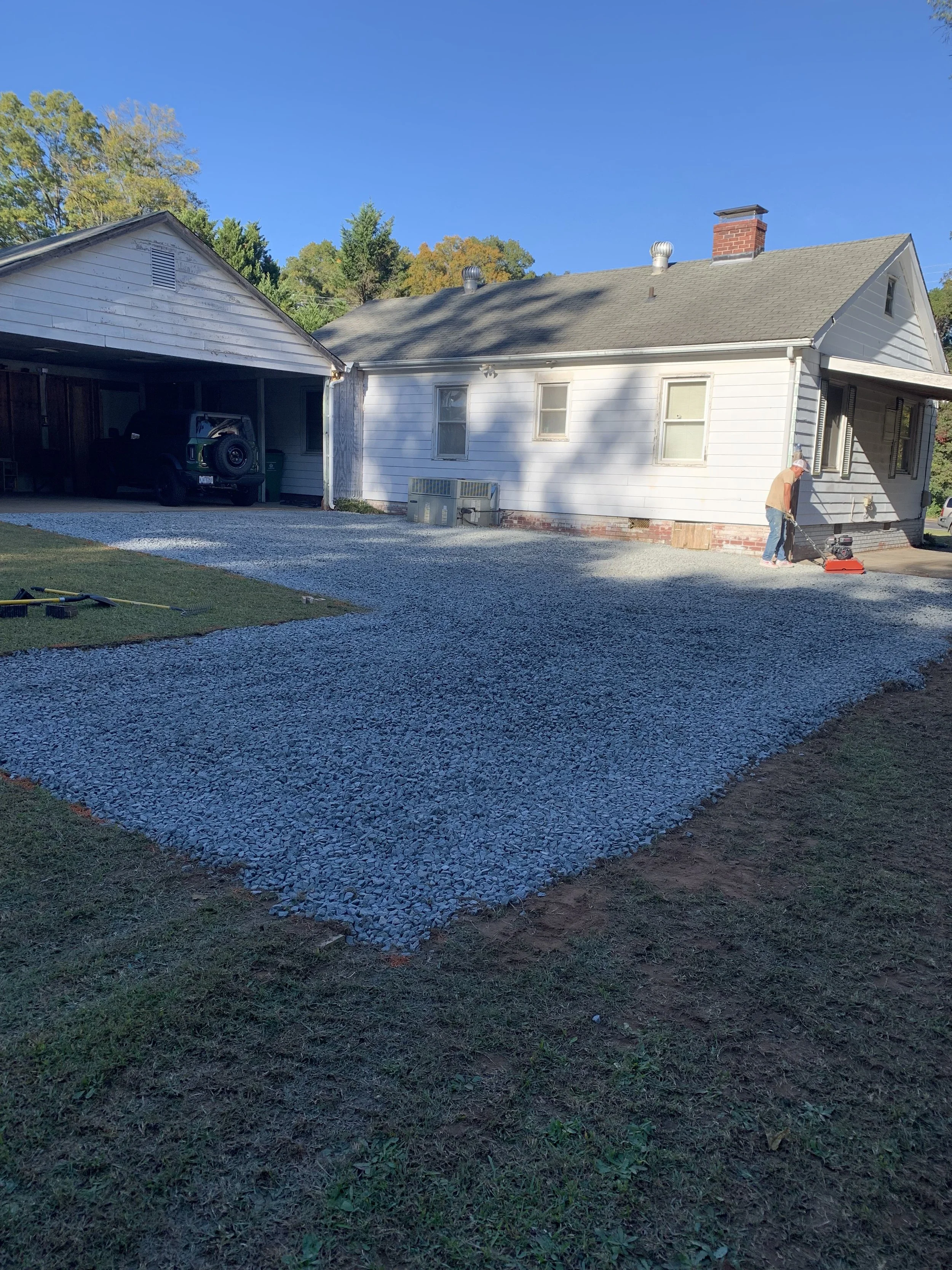 A house with a gravel driveway being prepared, with a person using a compactor, and a covered carport.