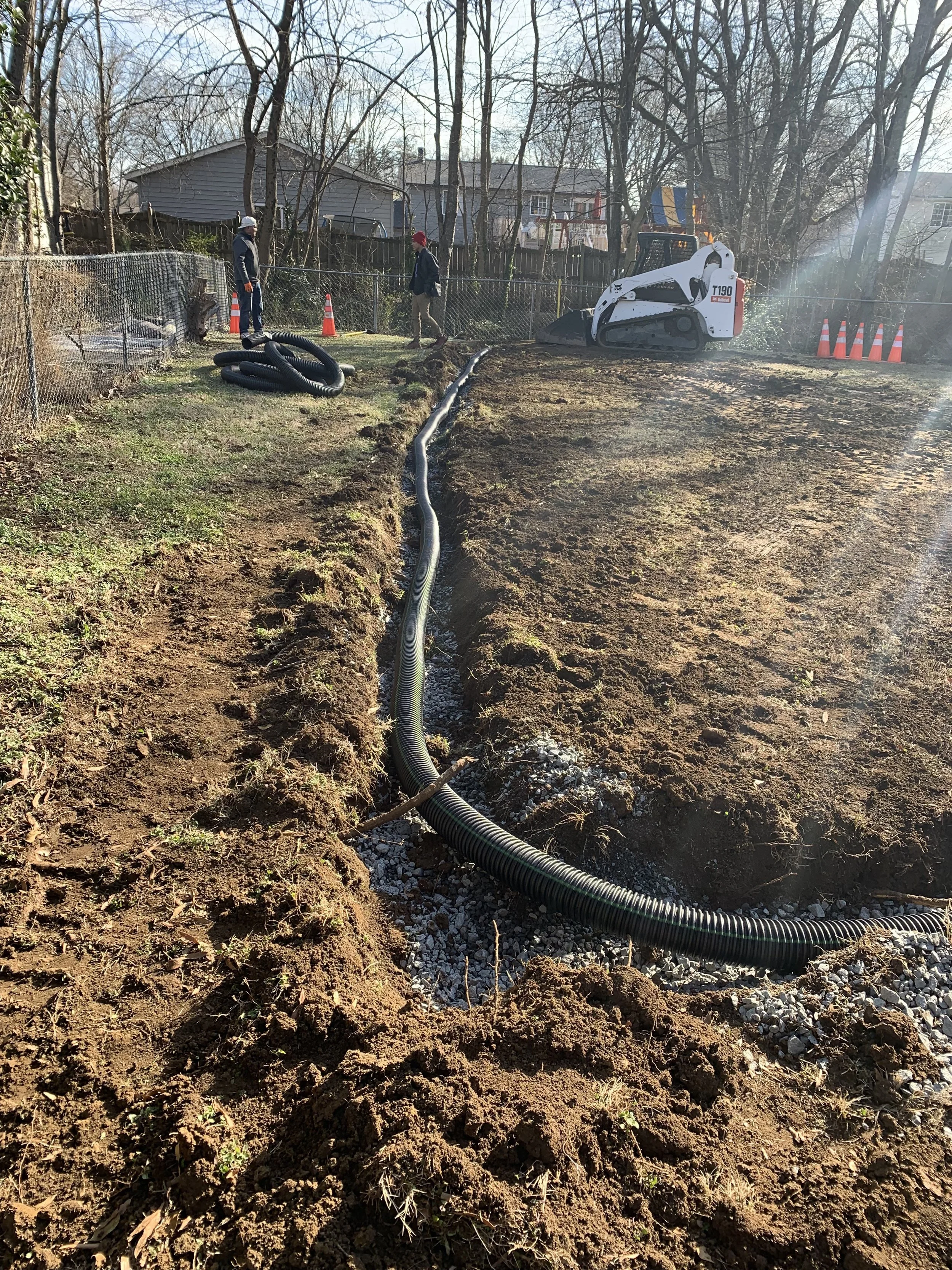 Construction workers installing underground piping with black corrugated tubing in a backyard, surrounded by soil and gravel, with a small excavator nearby.