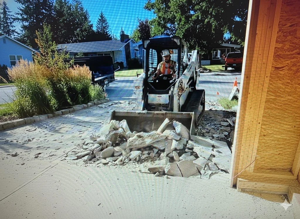 A construction worker operating a small skid-steer loader with a bucket attached, clearing debris outside a residential property, with houses, trees, and a parked pickup truck visible in the background.