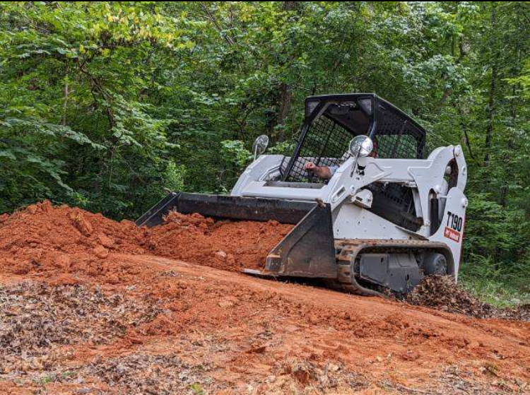 A white compact track loader moving reddish dirt on a construction site surrounded by green trees.