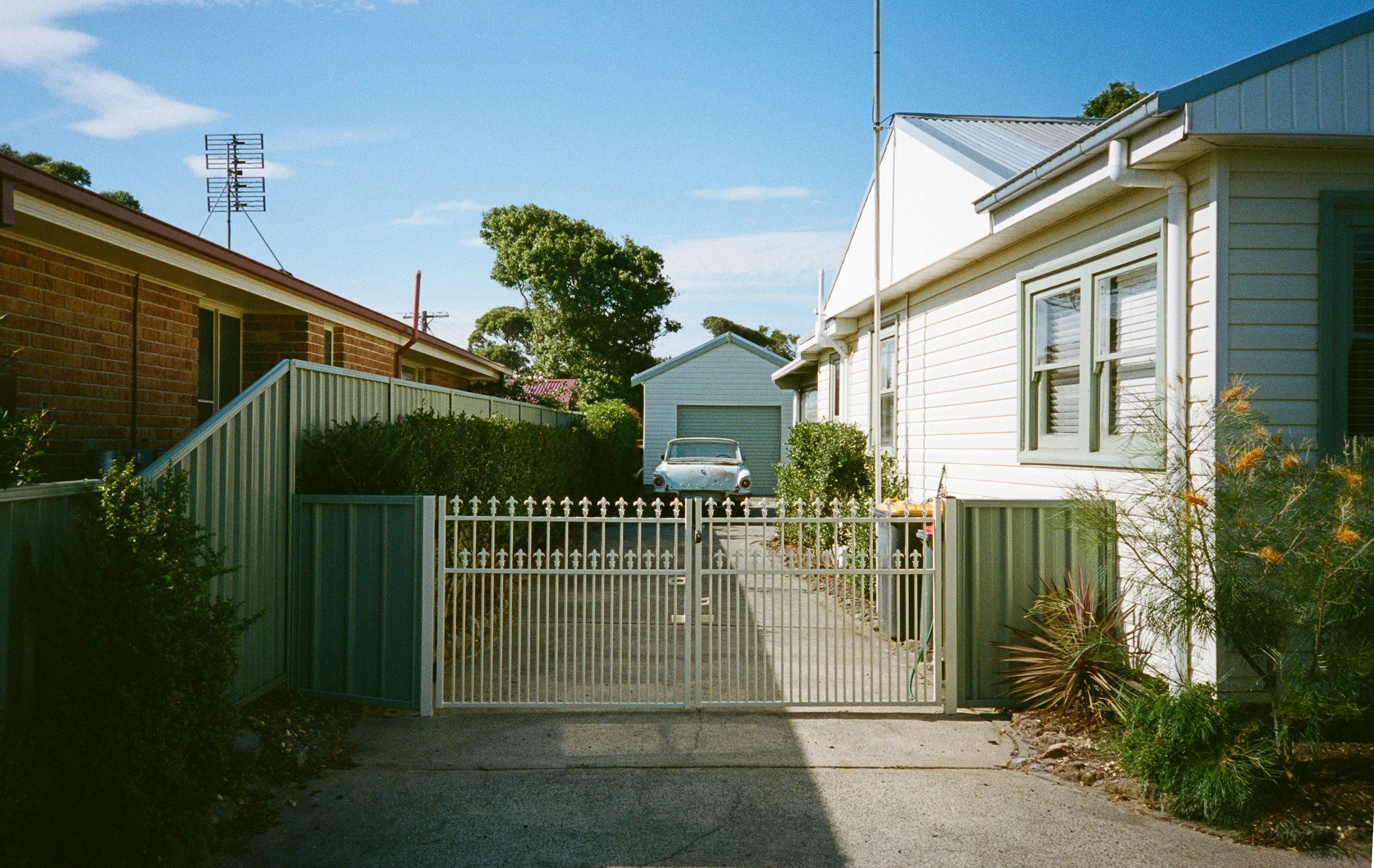 A driveway gated by a metal fence with a vintage car, possibly an old racing model, parked in front of a white garage in a suburban neighborhood. There are houses on either side and trees in the background under a blue sky.