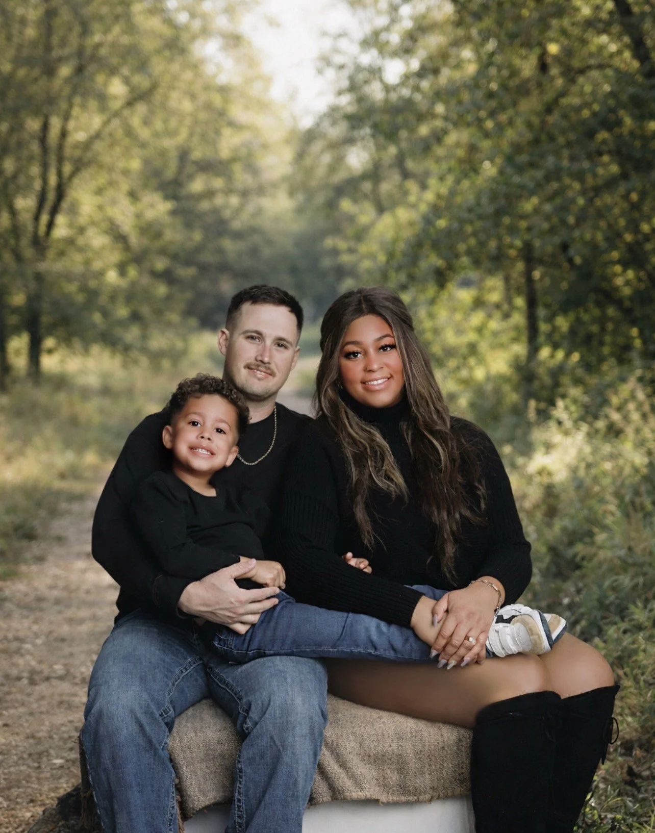 A family of three sitting on a bench outdoors in a forest setting. The man has short dark hair and a light beard, wearing a black shirt and blue jeans. The woman has long wavy brown hair, wearing a black turtleneck and black boots. The child, sitting on the woman's lap, has short curly hair and is smiling, wearing a black shirt and sneakers. All are smiling, with a backdrop of trees and greenery.