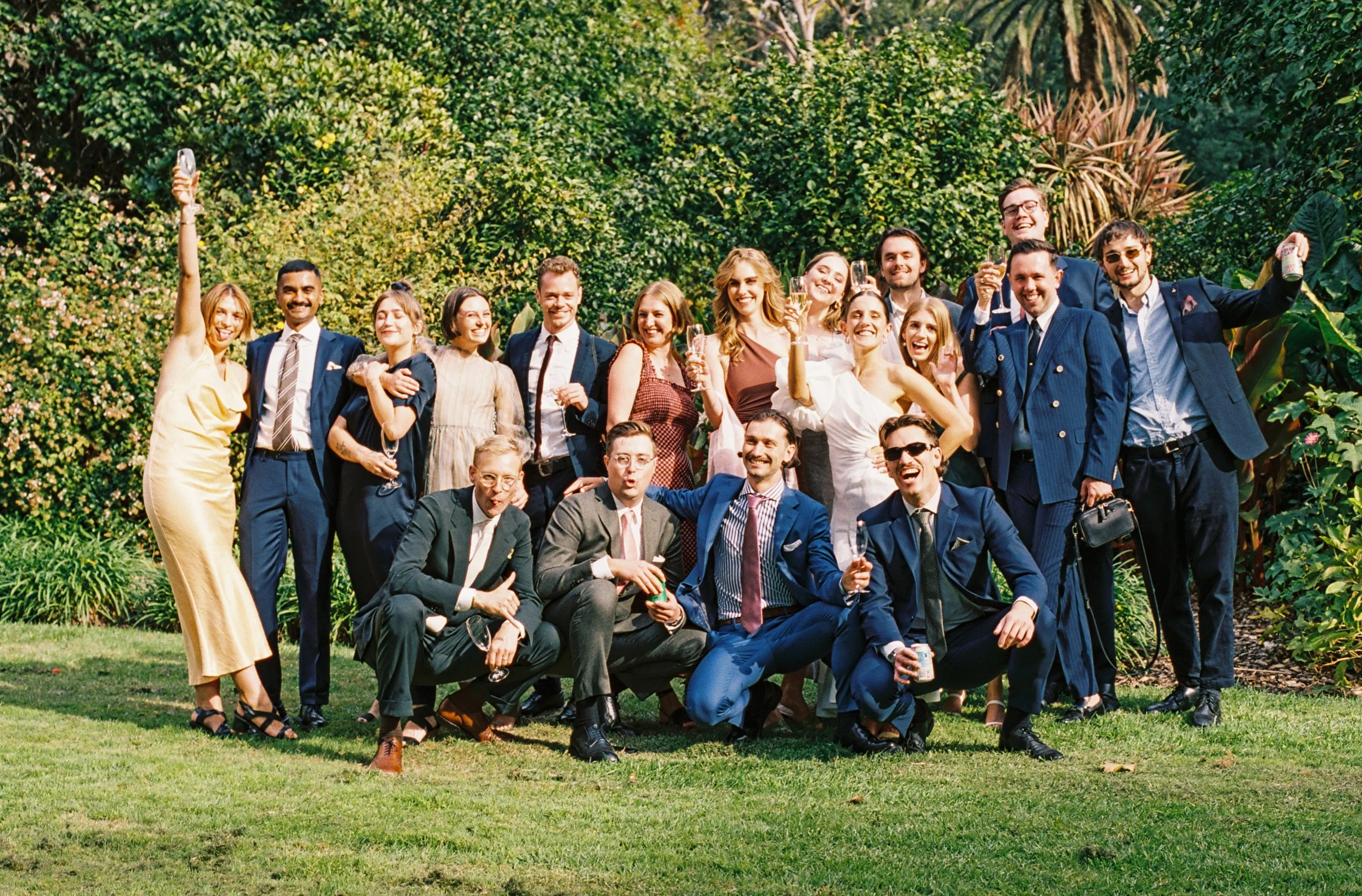 Group of people in formal attire celebrating outdoors in a lush garden, some holding drinks and smiling.