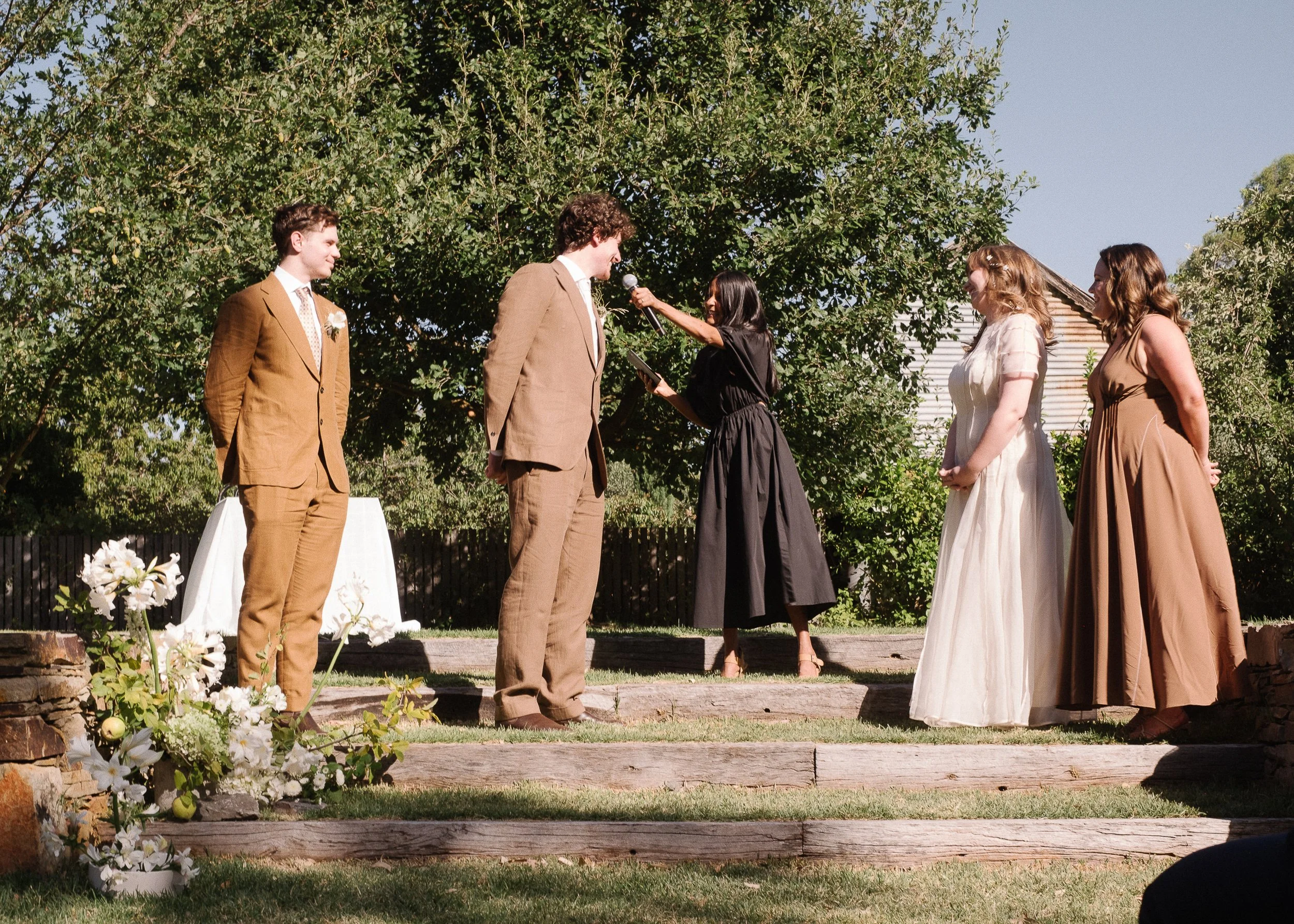 A wedding ceremony outdoors with five people standing on wooden steps. The groom and bride stand in the middle, with the groom reading vows and the bride holding a microphone and reading. Two women in dresses and two men in suits watch. Flowers and greenery are visible, with a large tree and house in the background.
