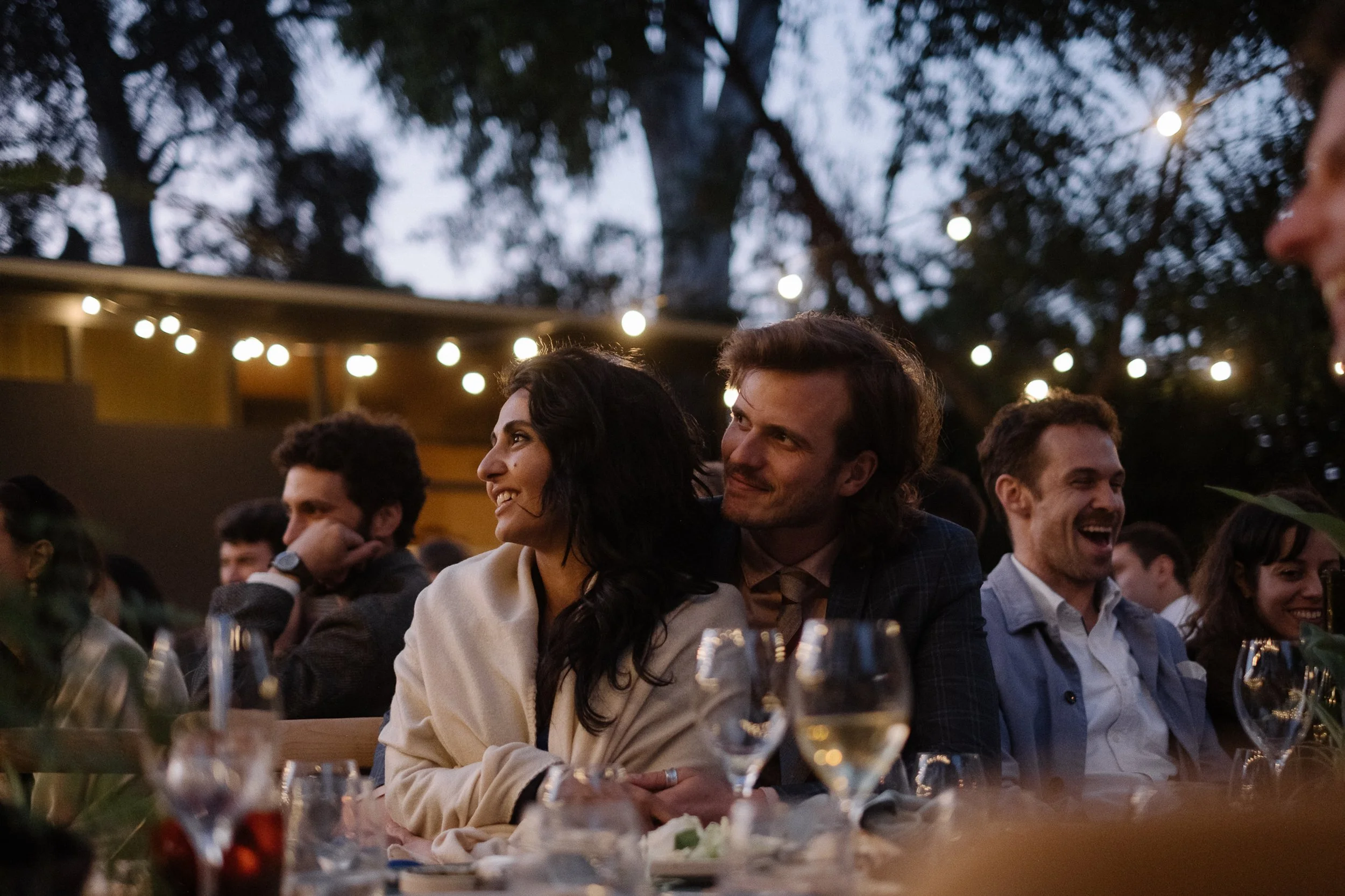 People sitting at an outdoor event at dusk, smiling and enjoying each other's company, with string lights hanging above.